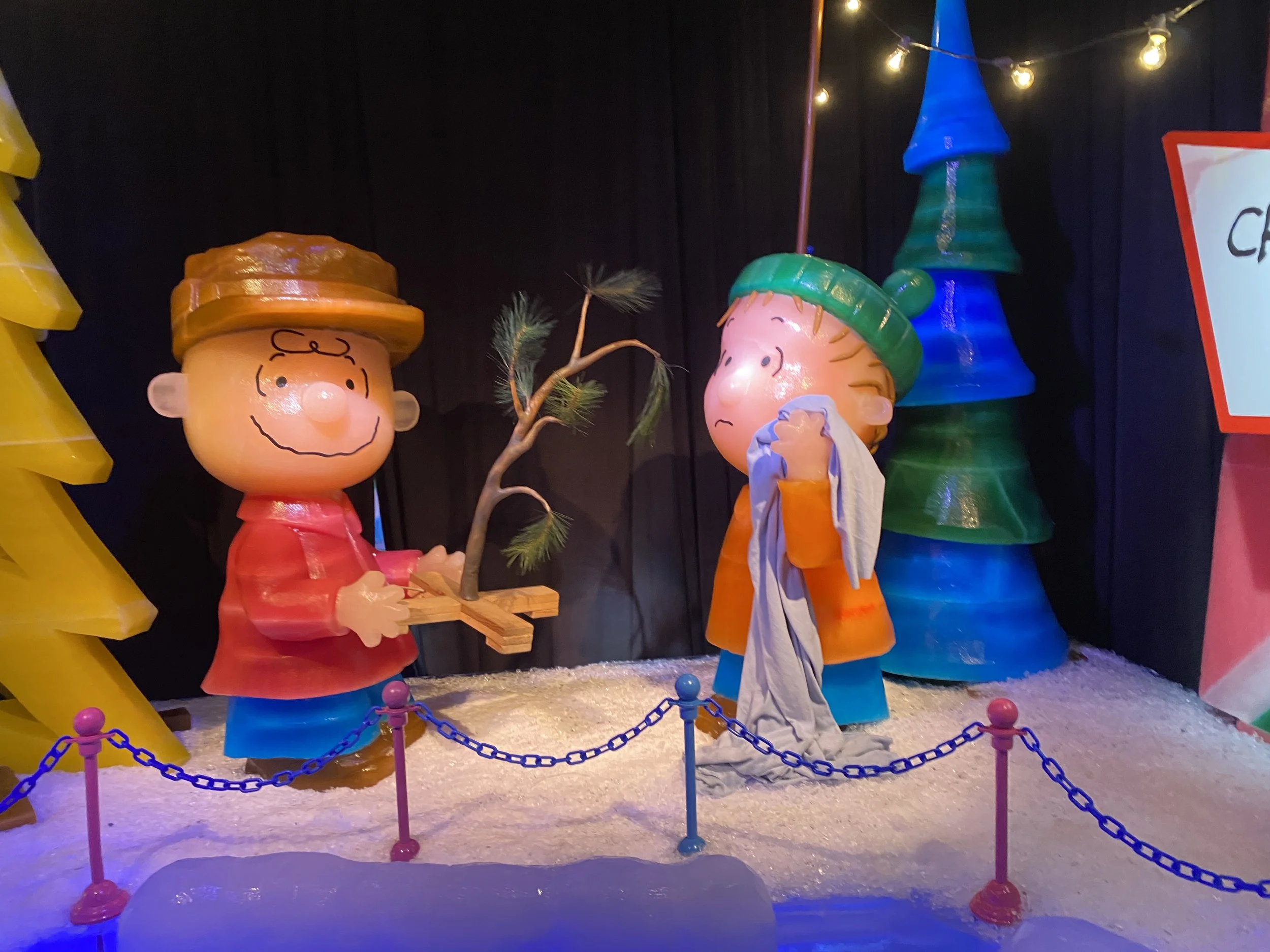 Colorful display of Charlie Brown and Lucy Van Pelt Christmas decorations from Peanuts, with bright lights and a small pine tree, enclosed by a blue chain barrier.