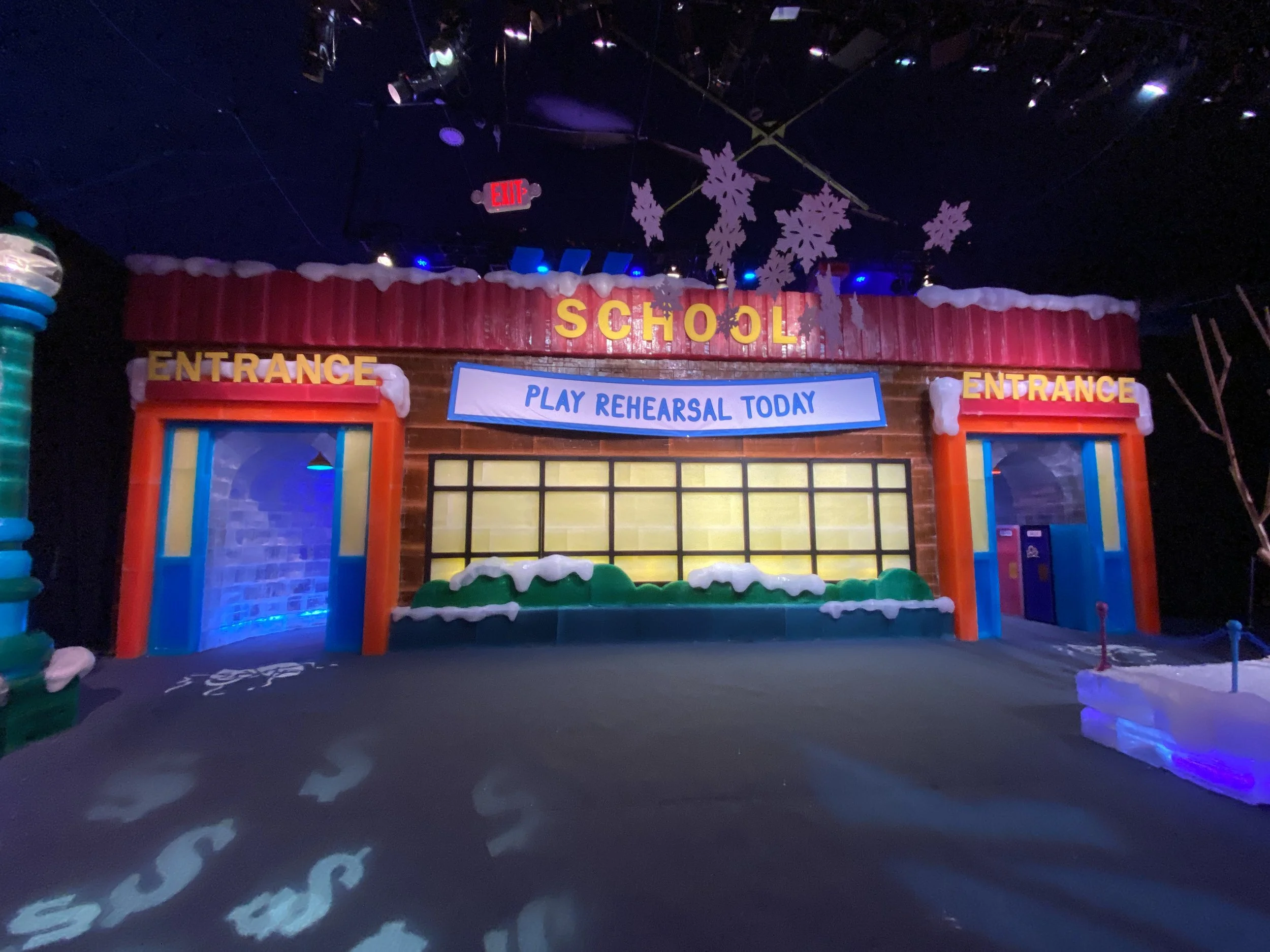 Decorated indoor space with a colorful facade marked 'School', snowflake decorations hanging from the ceiling, and lights creating patterns on the floor. A banner says 'Play Rehearsal Today'.