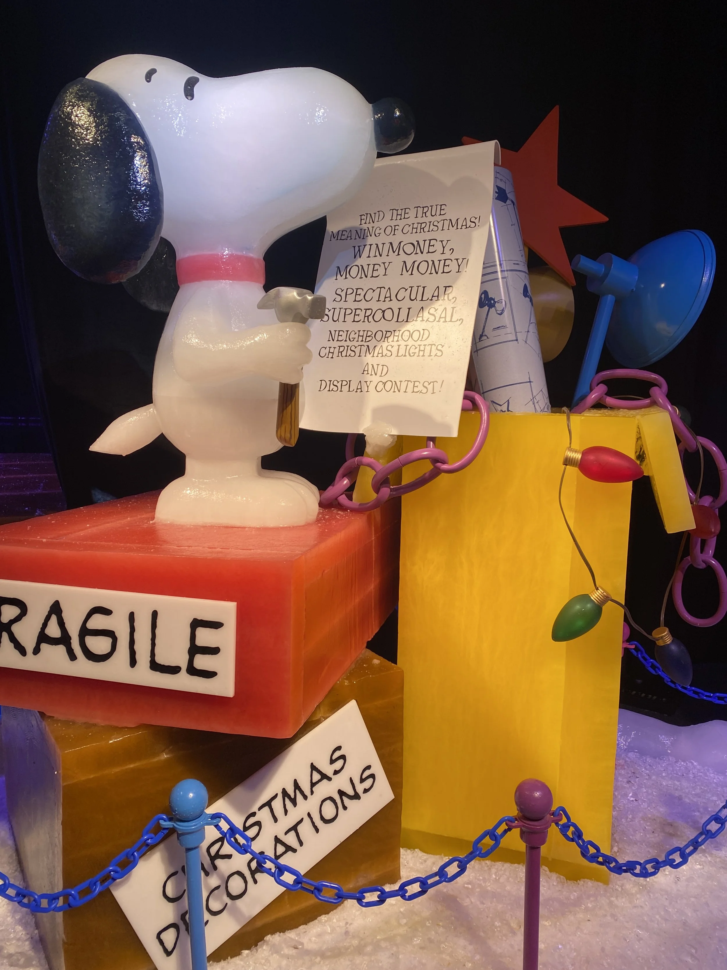 Decorative Snoopy figurine holding a sign with Christmas-related text, surrounded by colorful holiday lights and chains, with a yellow block and another sign, and artificial snow on the ground.