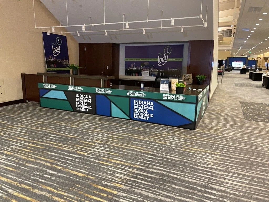 An information booth for the Indiana Global Economic Summit 2024 inside an airport or conference center, with a purple backdrop and a small counter with plants and signs.