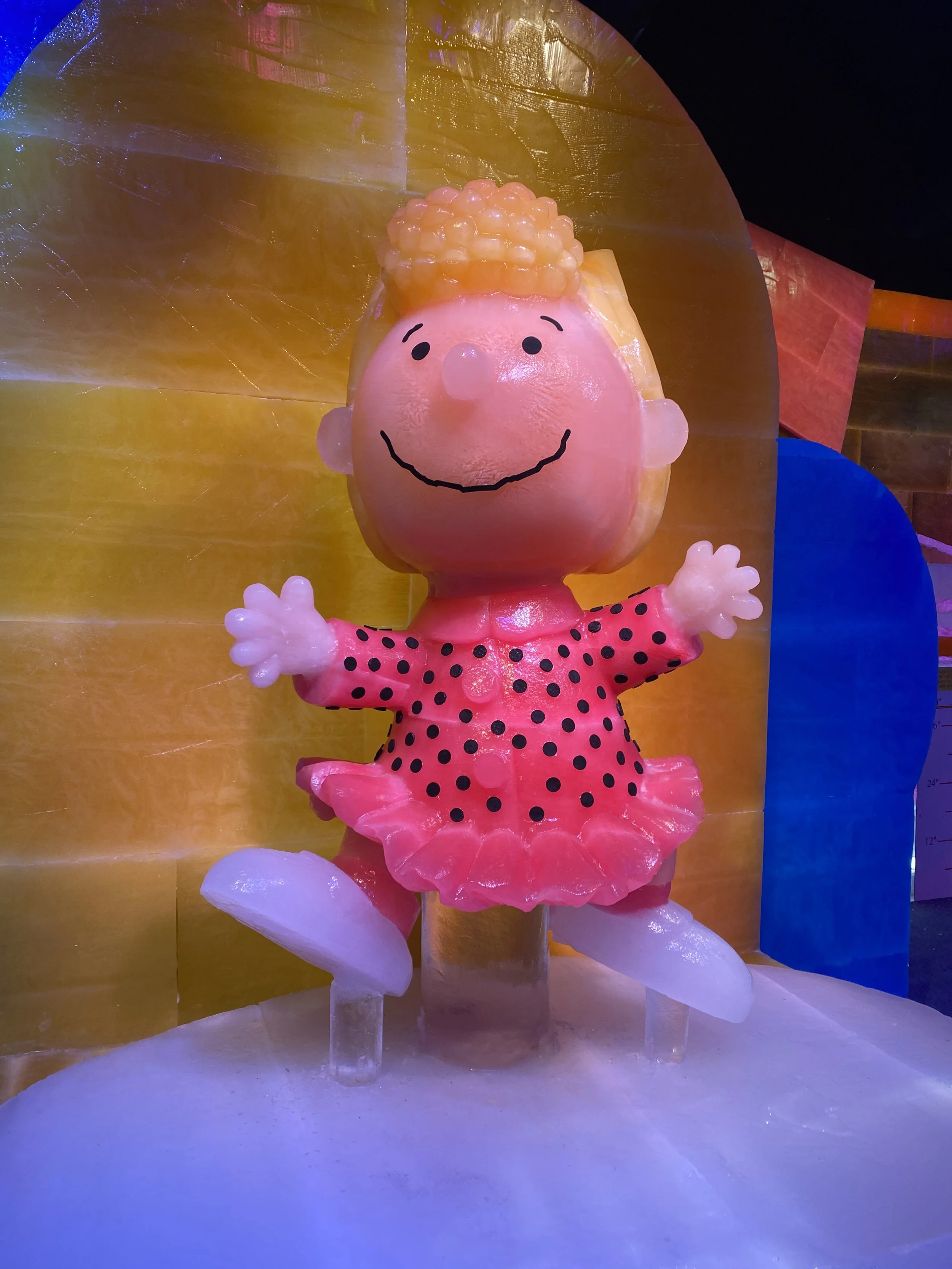 Ice sculpture of a cartoon girl with a pink face, red and black polka-dotted dress, smiling face with drawn eyes, nose, and mouth, with yellow curly hair made of ice and a yellow background.
