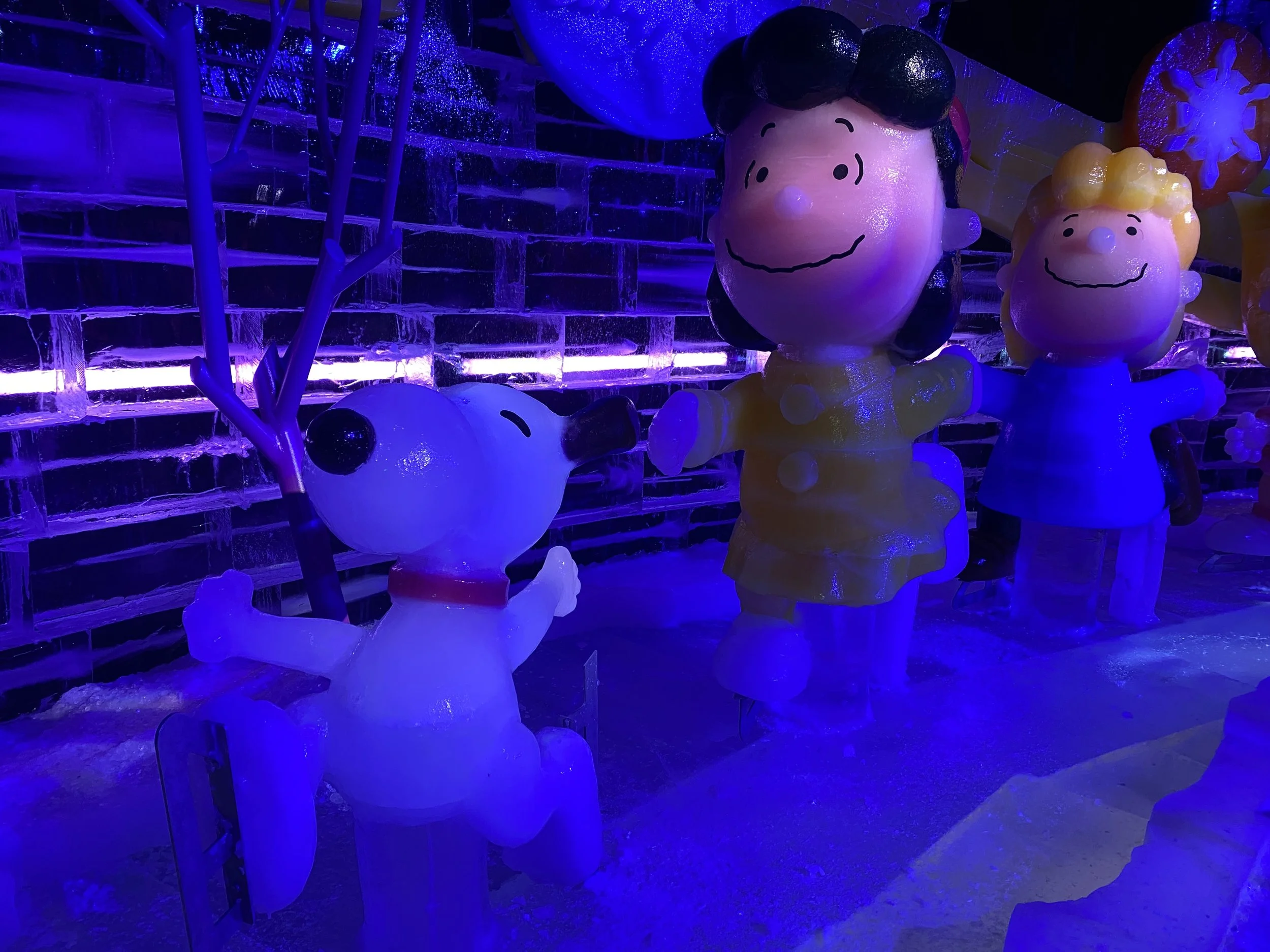 Ice sculpture display featuring characters from Peanuts, including Charlie Brown, Lucy, and Snoopy, illuminated by vibrant blue and purple lights.