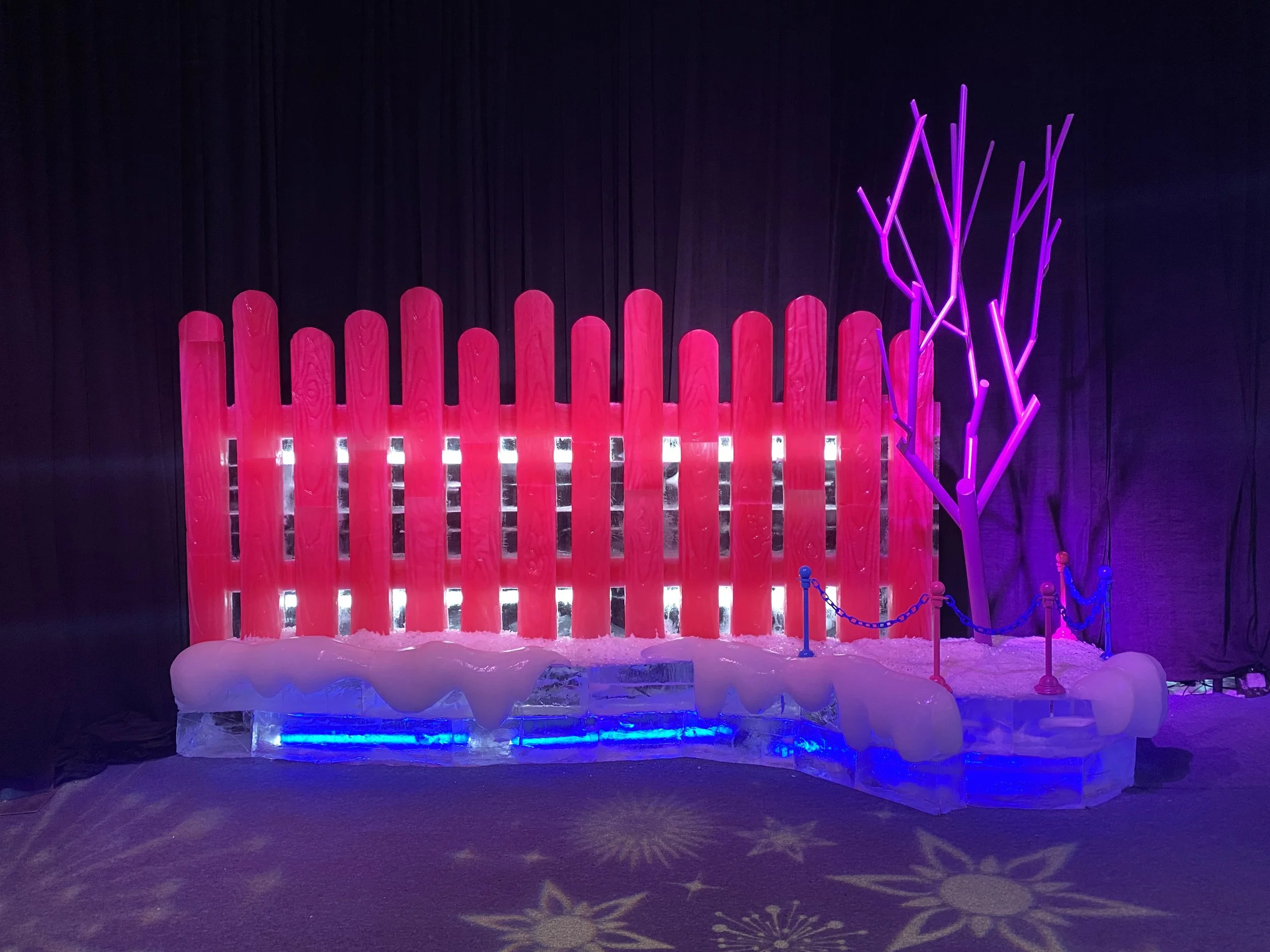 Colorful ice sculpture display with pink fences, purple neon tree, and blue-colored base lighting, set against a dark background.