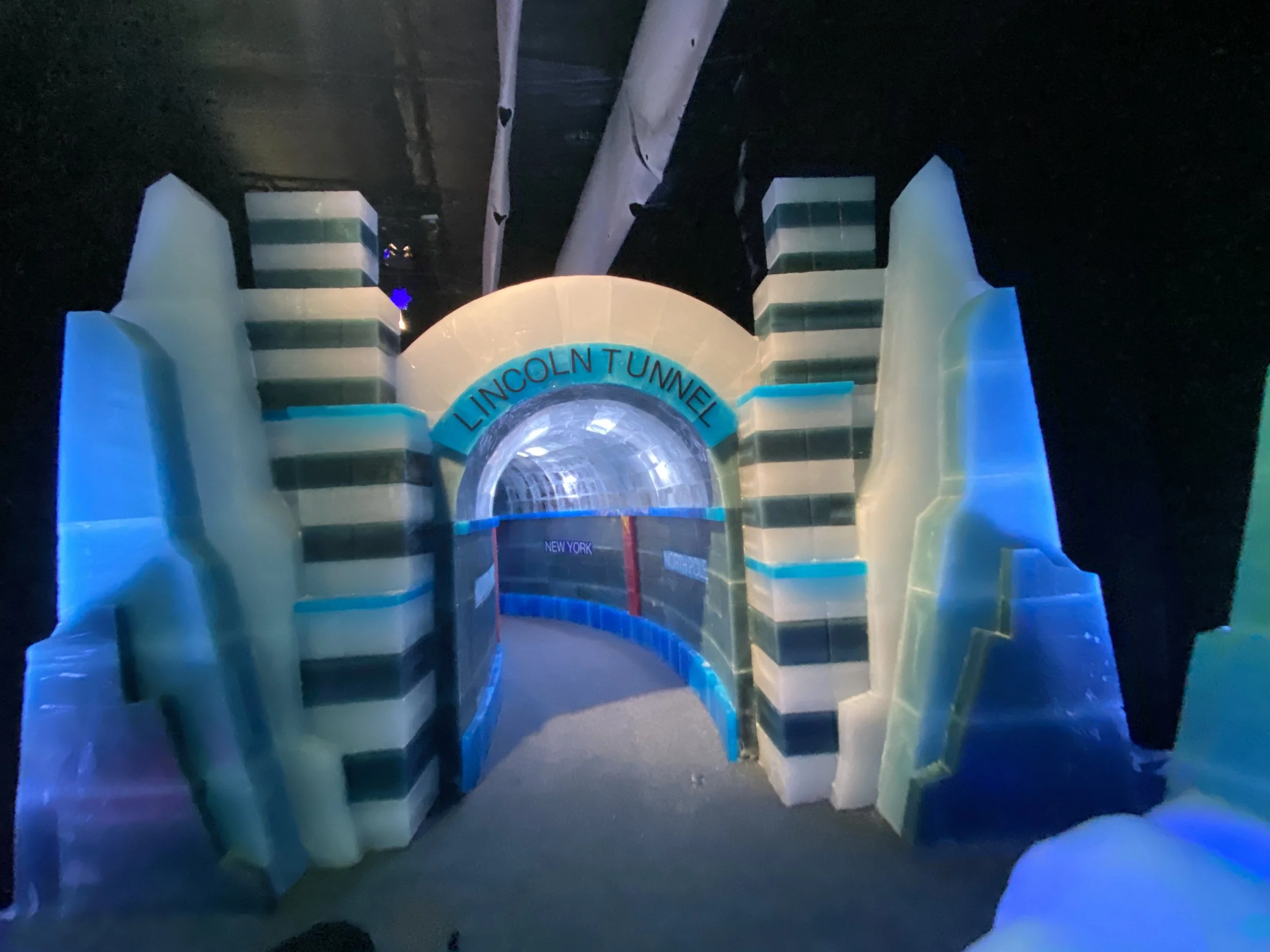 Ice tunnel exhibit with curved, striped walls illuminated by blue and green lights, labeled Lincoln Tunnel and New York.