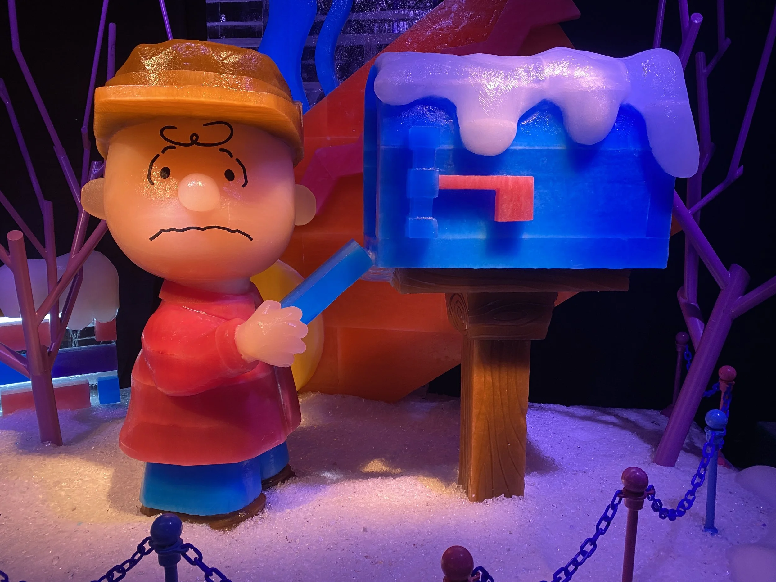 A colorful illuminated display of cartoon characters, including a sad-looking child's face with an orange hat and a white dripping object, possibly snow or ice, in a dark setting with purple and pink accents.