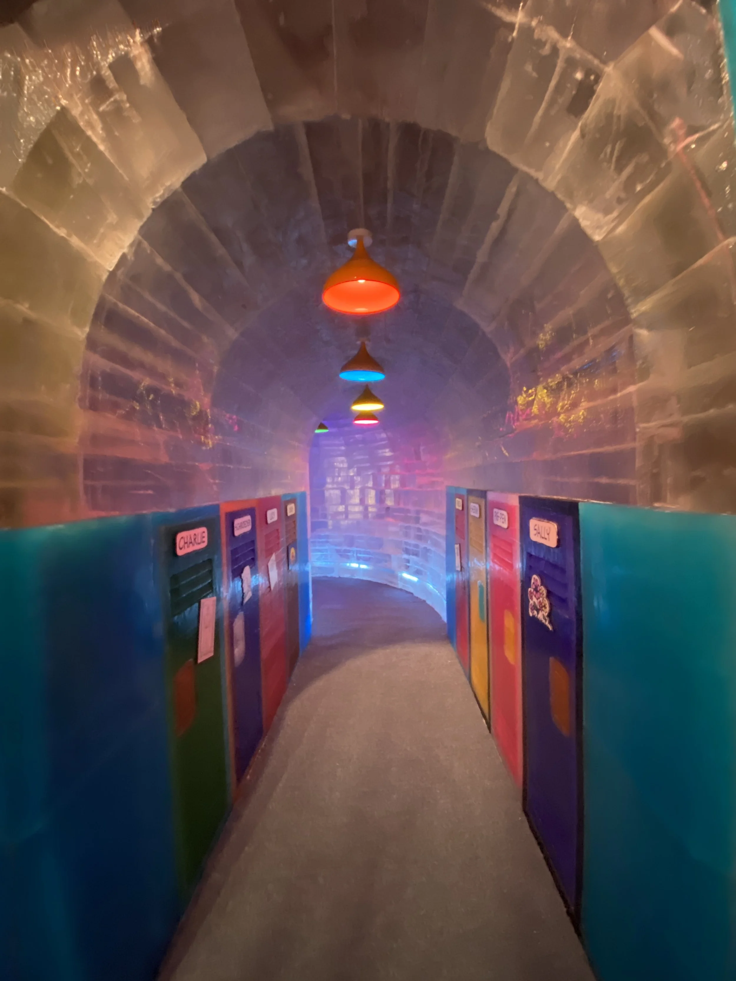 Colorful hallway with a brick arched ceiling, decorated with hanging pendant lights in red, yellow, blue, and green colors, and walls lined with colorful lockers that have labels on them.