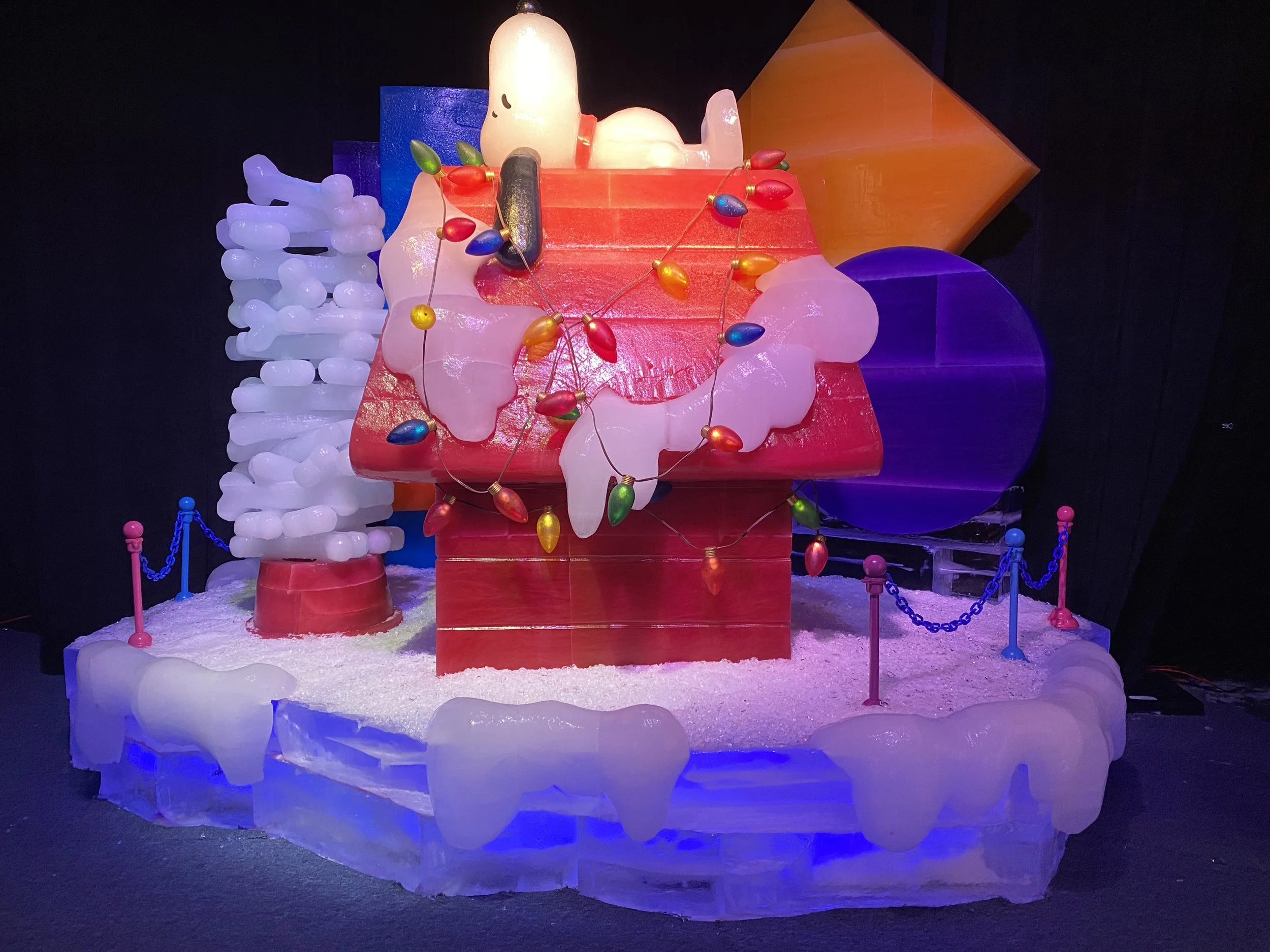 Ice sculpture display resembling a cartoon character, decorated with colorful string lights, set in a snowy environment with a dark background.