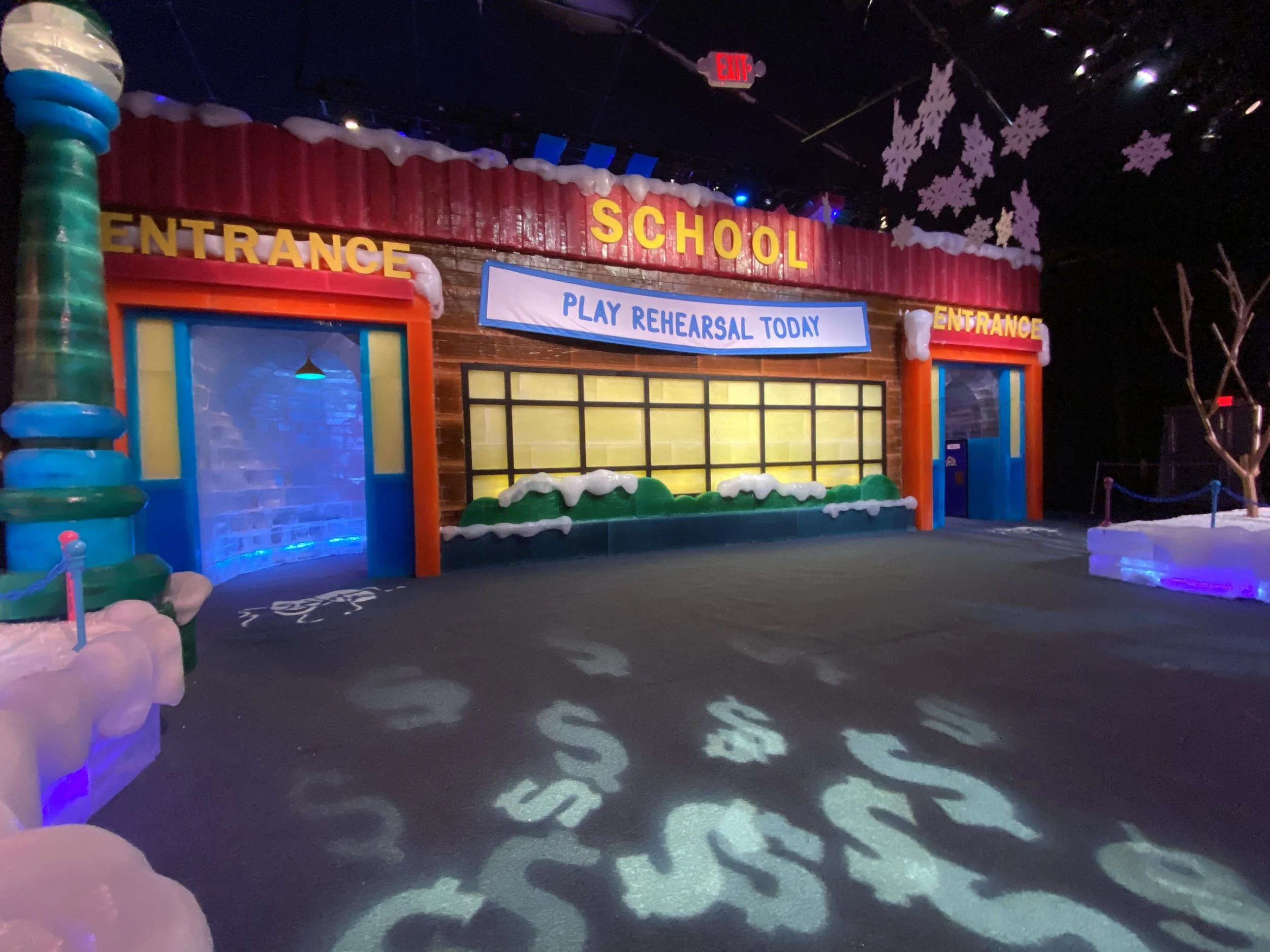 Decorated indoor display with a sign that says 'Play Rehearsal Today' and large yellow and red words 'ENTRANCE' and 'SCHOOL'; themed with snow, winter trees, and festive decorations.