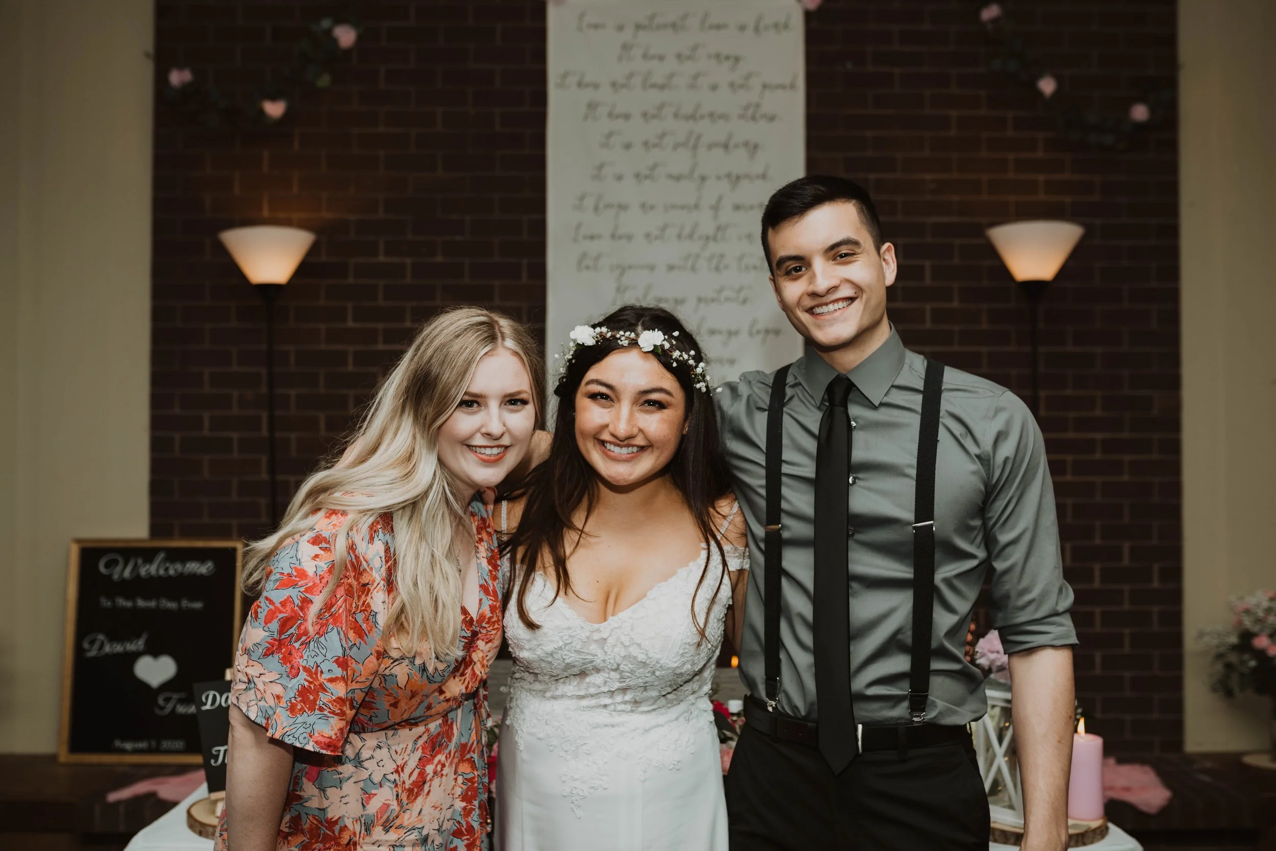 Three friends smiling at a wedding reception, with the bride in a white dress and floral crown in the center, flanked by a woman in a colorful dress on the left and a man in a gray shirt with suspenders on the right, standing in front of a brick wall