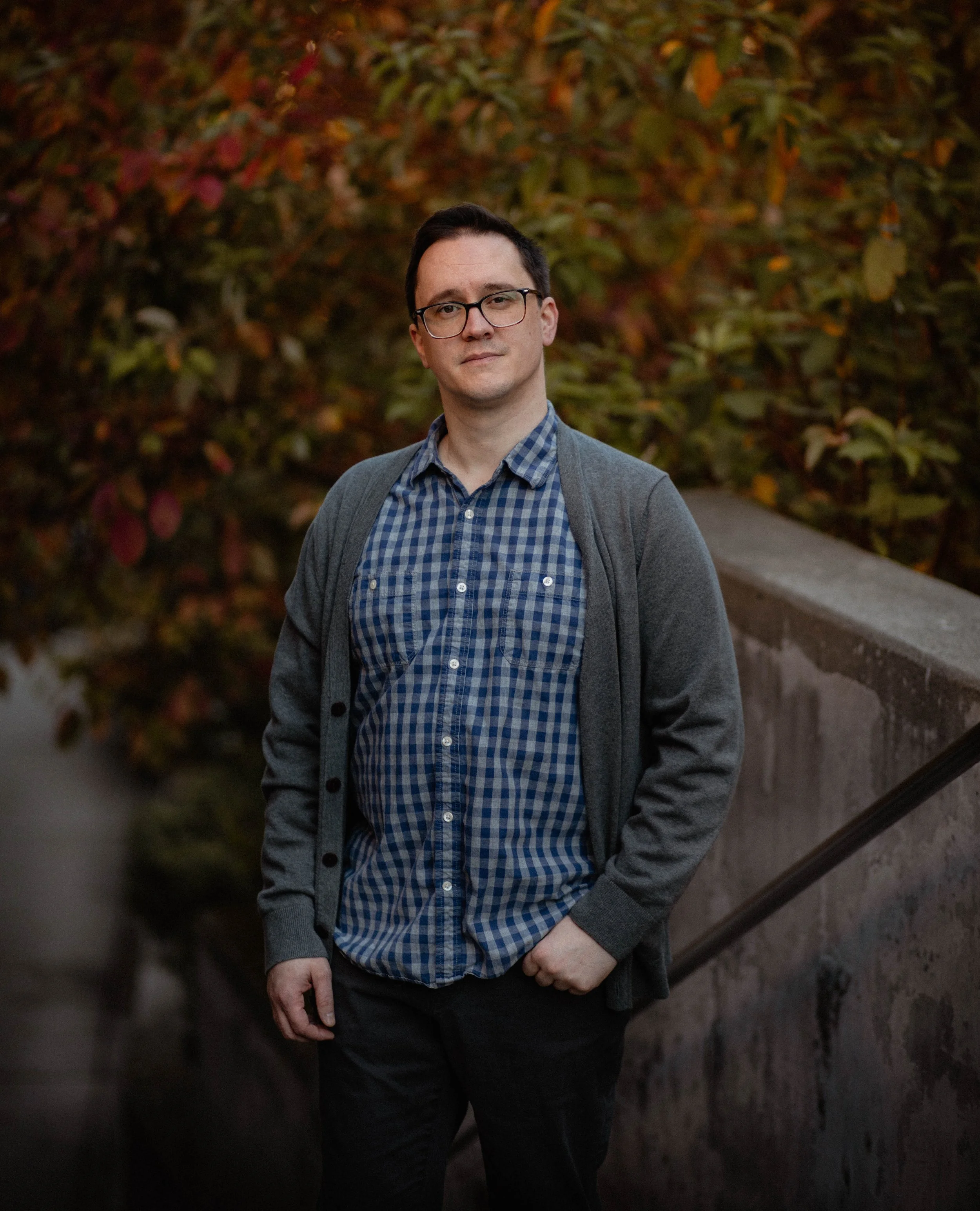 A man with glasses wearing a blue checkered shirt and gray cardigan standing outdoors with fall foliage in the background. Seattle professional head shot photography