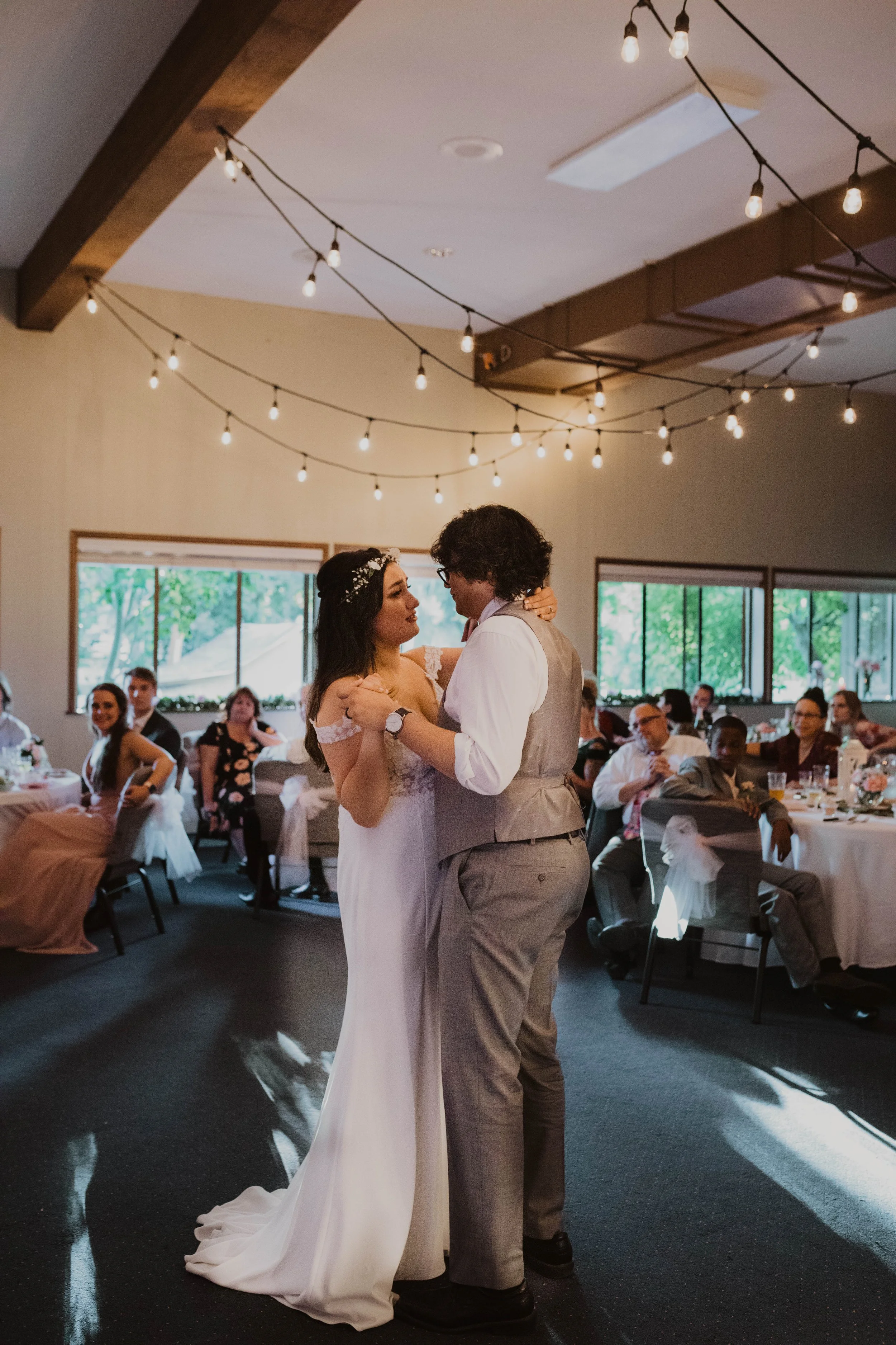 A bride and groom share their first dance at a wedding reception, with seated guests watching in the background and string lights hanging from the ceiling.Seattle, WA wedding photography. 