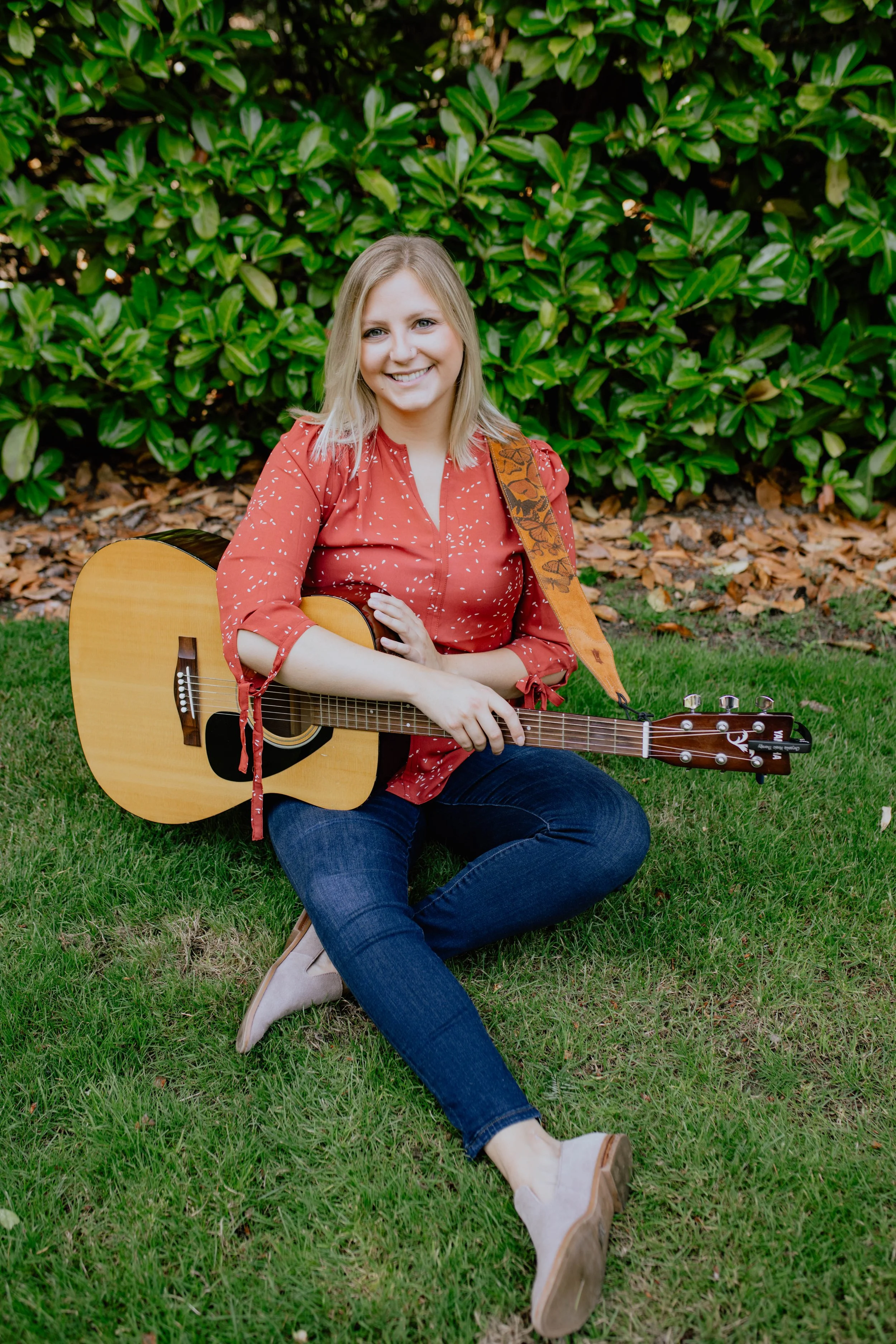 A woman with blonde hair sitting on grass, holding an acoustic guitar, smiling, wearing a red blouse with white patterns, blue jeans, and beige shoes, with green bushes and brown leaves in the background. Seattle professional head shot photography