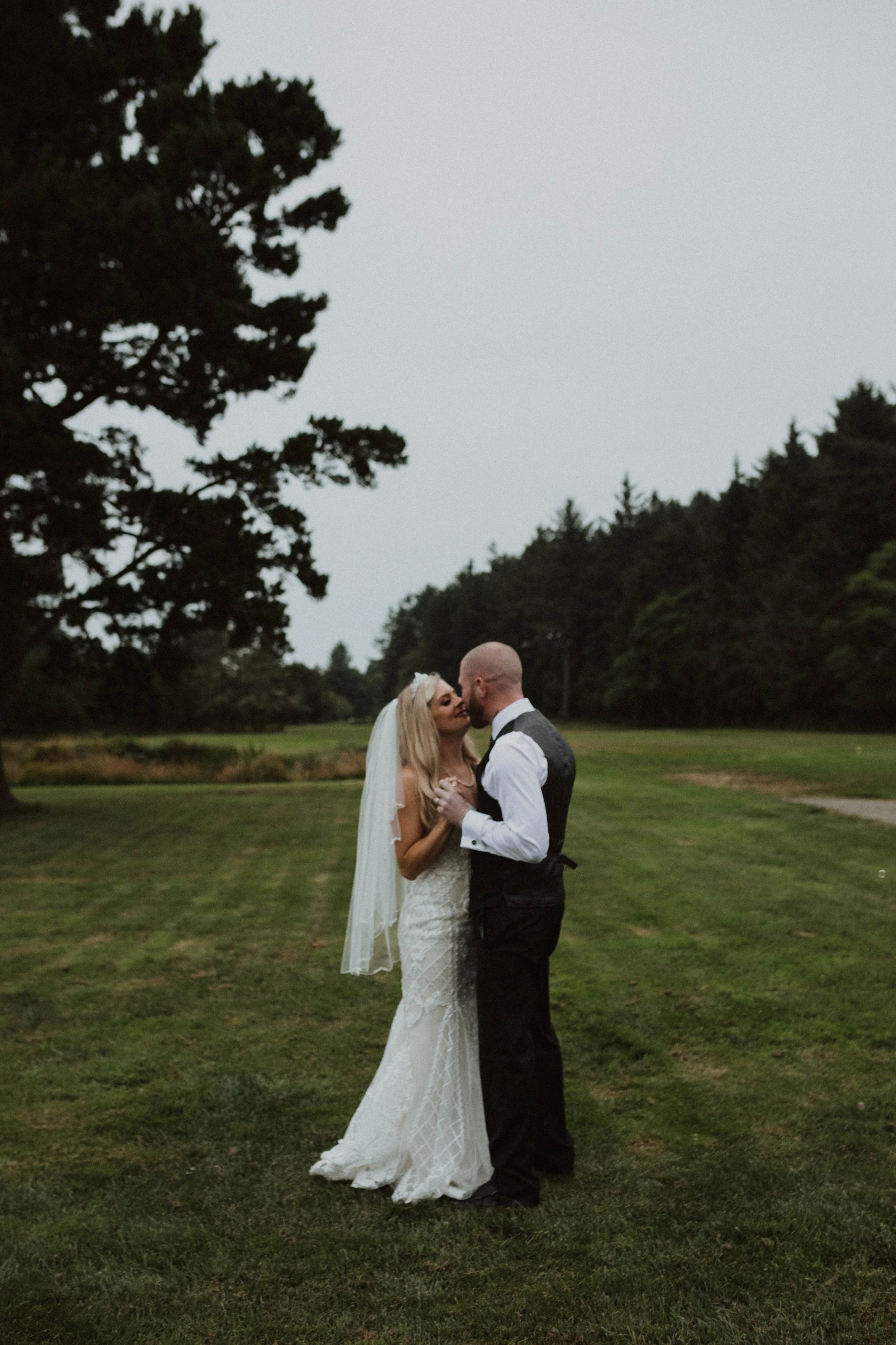 A bride and groom sharing a kiss outdoors on a grassy field, with trees and an overcast sky in the background. Long Beach, WA wedding photography.