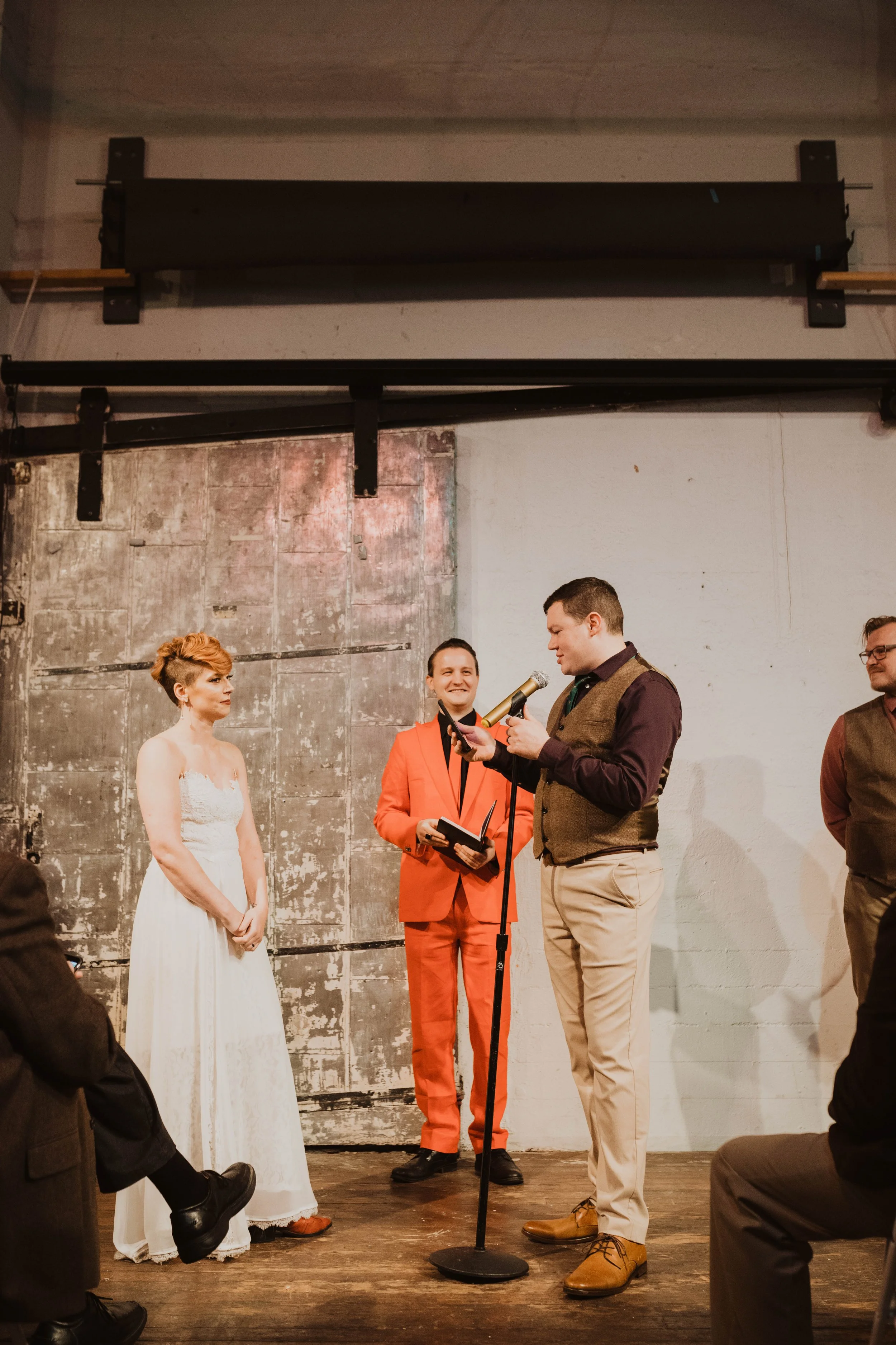 At a wedding ceremony, a bride in a white dress stands on the left, and a groom in a vest and beige pants holds a microphone and a piece of paper, speaking. An officiant in a bright red suit stands behind them, smiling.