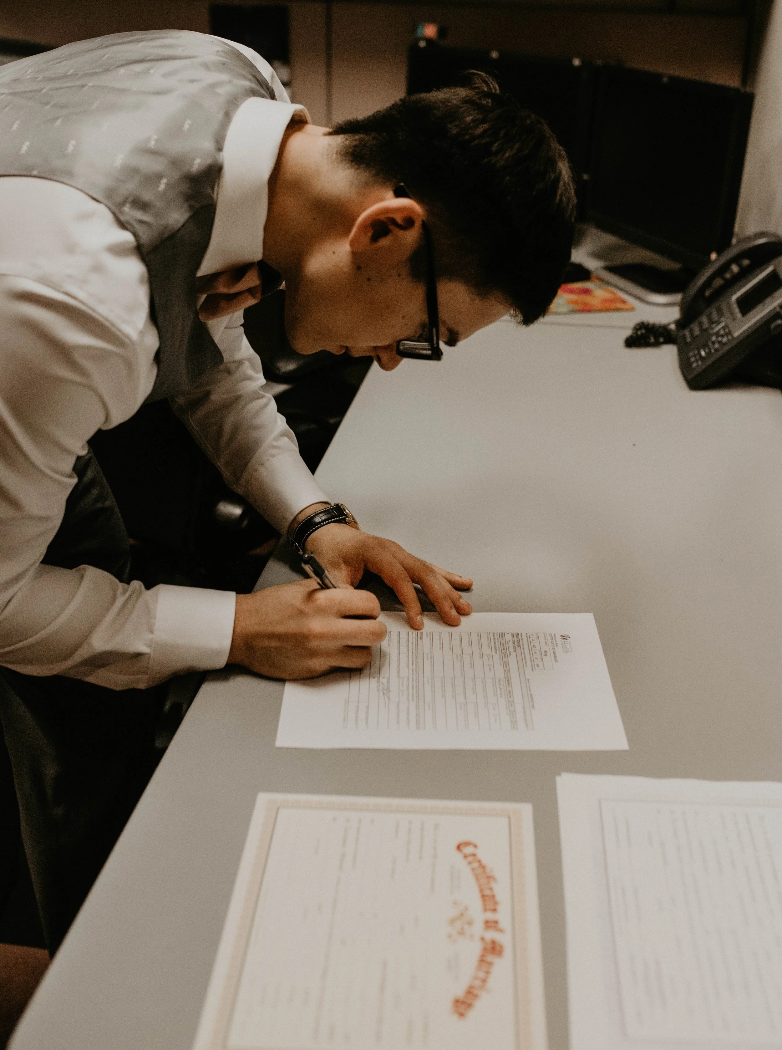 A man wearing glasses and a white shirt signs a document on a gray table in an office setting. Seattle, WA wedding photography.
