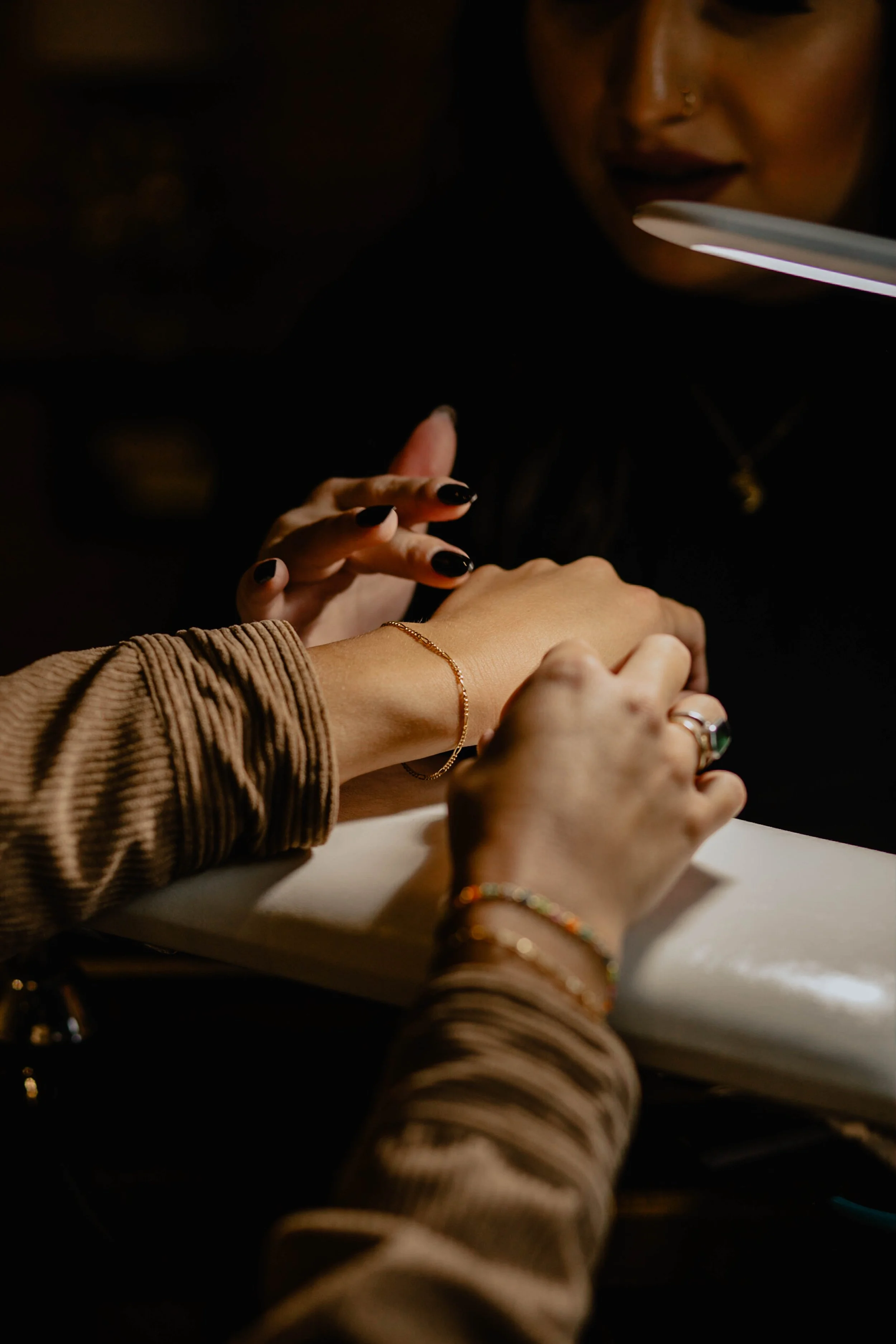 Close-up of two women practicing a manicure, one woman painting the other's nails in a dimly lit setting, with jewelry and dark clothing. Seattle professional head shot photography
