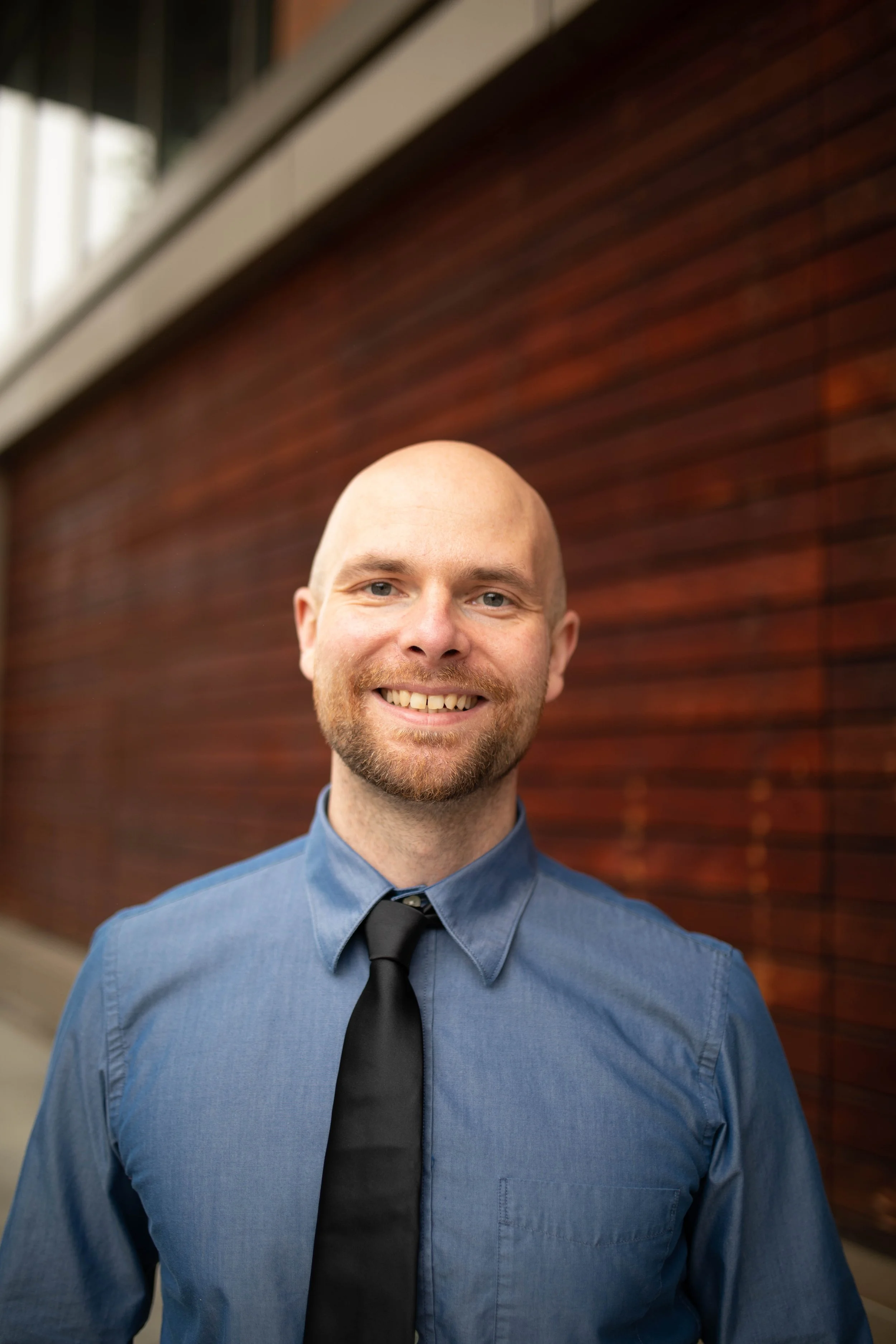 A smiling man with a beard and bald head wearing a blue shirt and a black tie, standing outdoors against a blurred red brick wall background. Seattle professional head shot photography