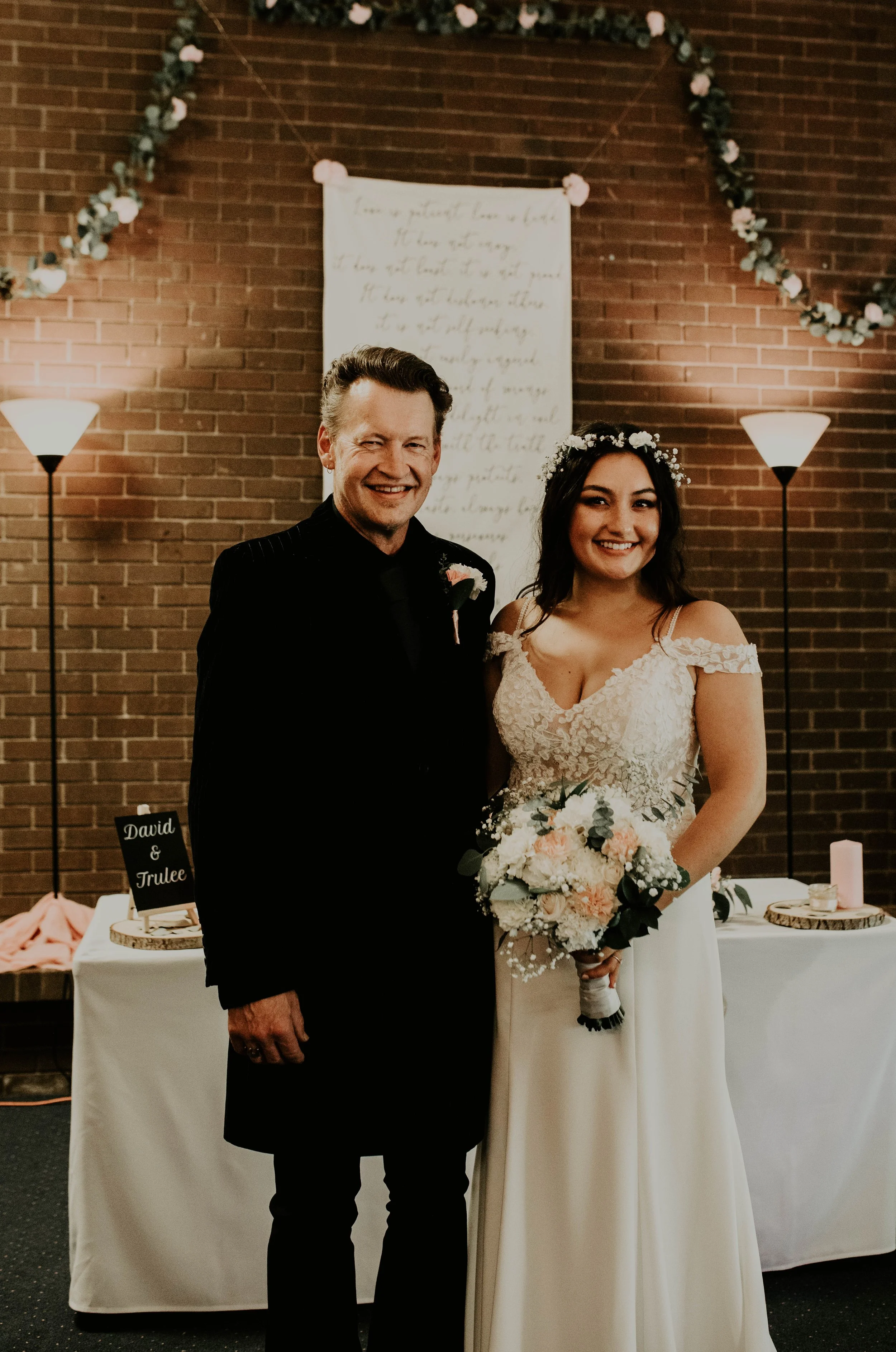 A bride and a man are standing together indoors during a wedding celebration. The bride is holding a bouquet of flowers, wearing a white lace wedding dress with spaghetti straps and a floral crown. The man, dressed in a black suit, is smiling and win