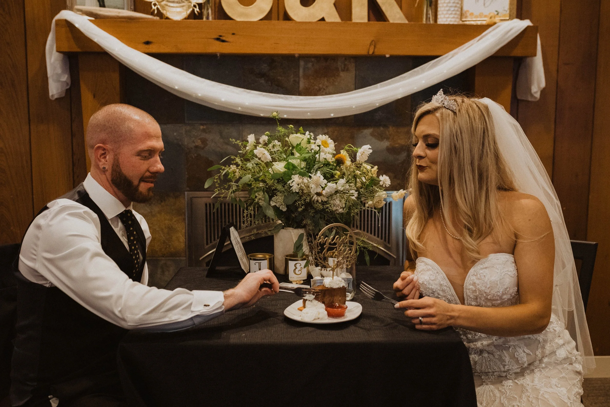 A bride and groom sitting at a table during their wedding reception, with a floral centerpiece and decorations in the background. Long Beach, WA wedding photography.