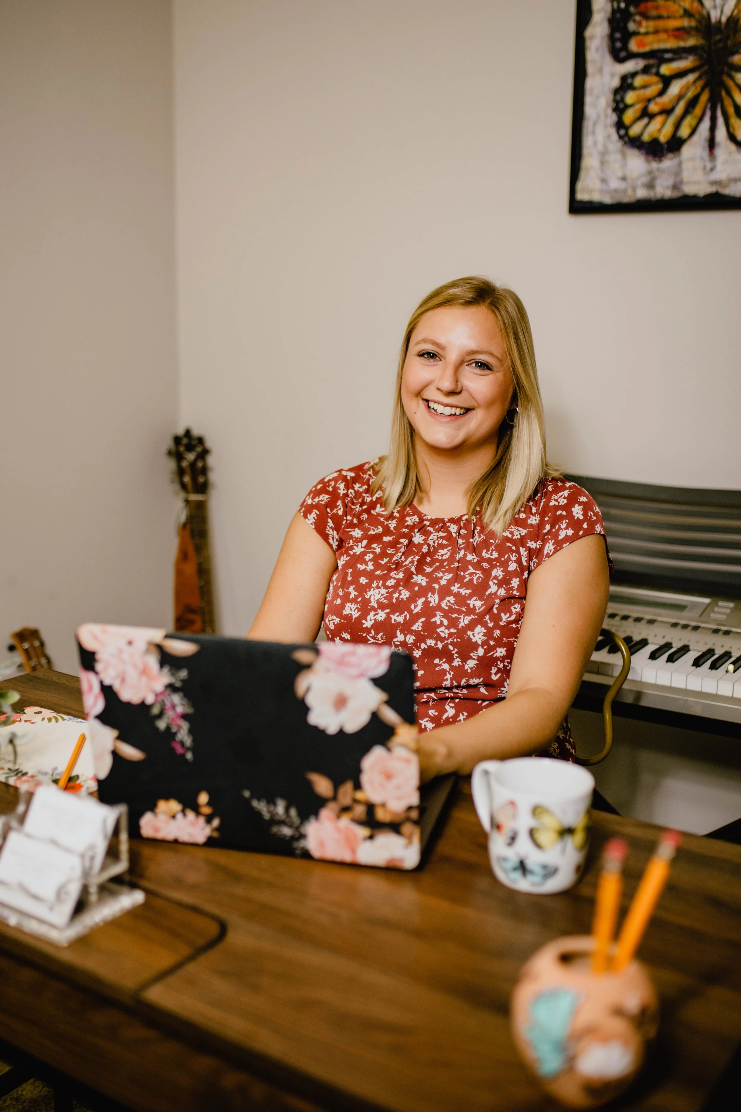 A woman with blonde hair wearing a red floral shirt sitting at a wooden table with a floral-patterned laptop, a mug with a butterfly design, and other stationery. There is a keyboard in the background and a butterfly artwork on the wall.