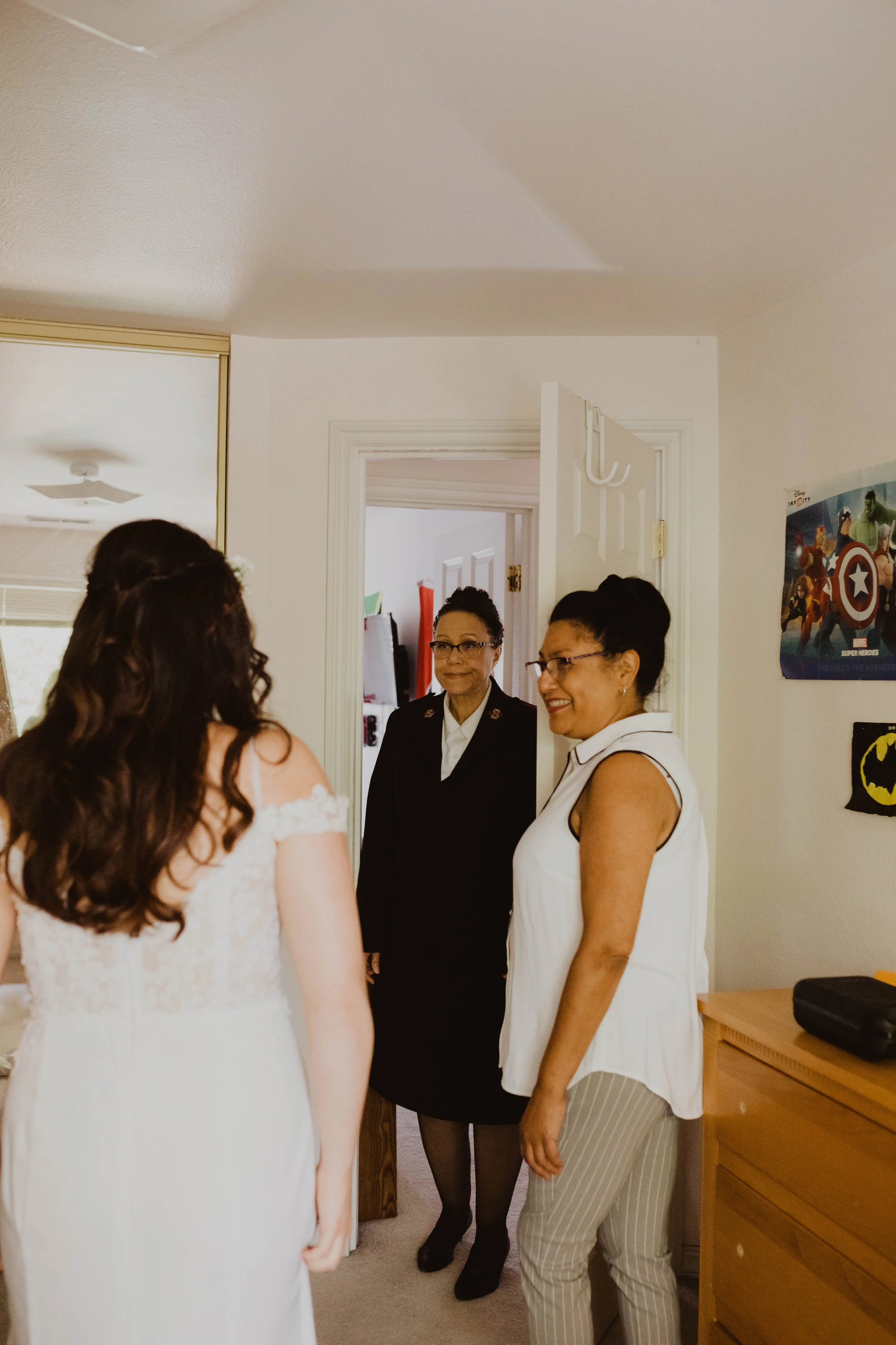 Three women in a room; one with long brown hair in a white dress, another in a dark uniform with glasses, and a third in a sleeveless white top with striped pants, all engaging in conversation. Seattle, WA wedding photography.