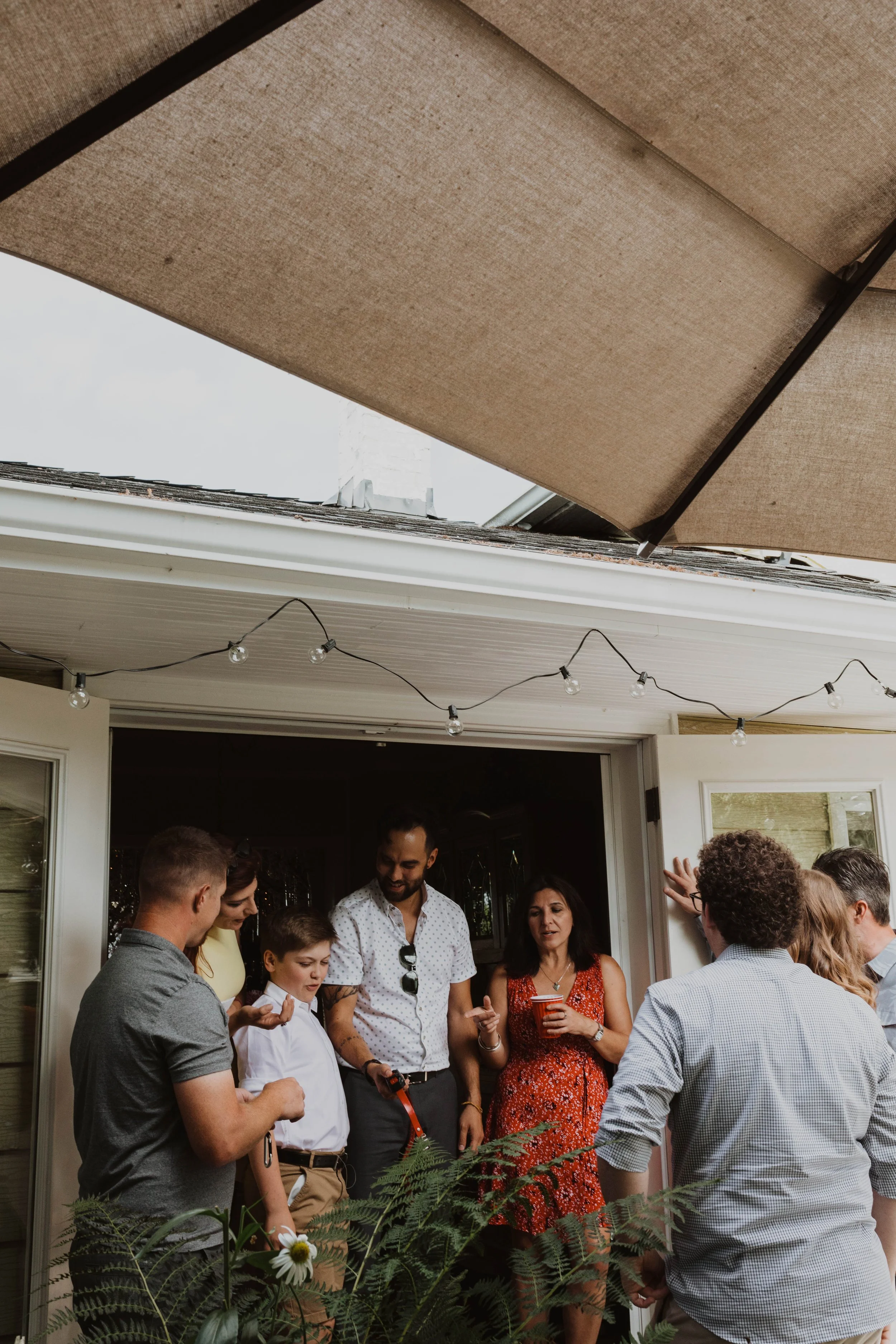 A group of people gathered on a porch, engaging in conversation during a backyard gathering. There is a man holding a red cup, a woman with a drink in her hand, and a young boy among others. String lights are hanging above, and a large patio umbrella