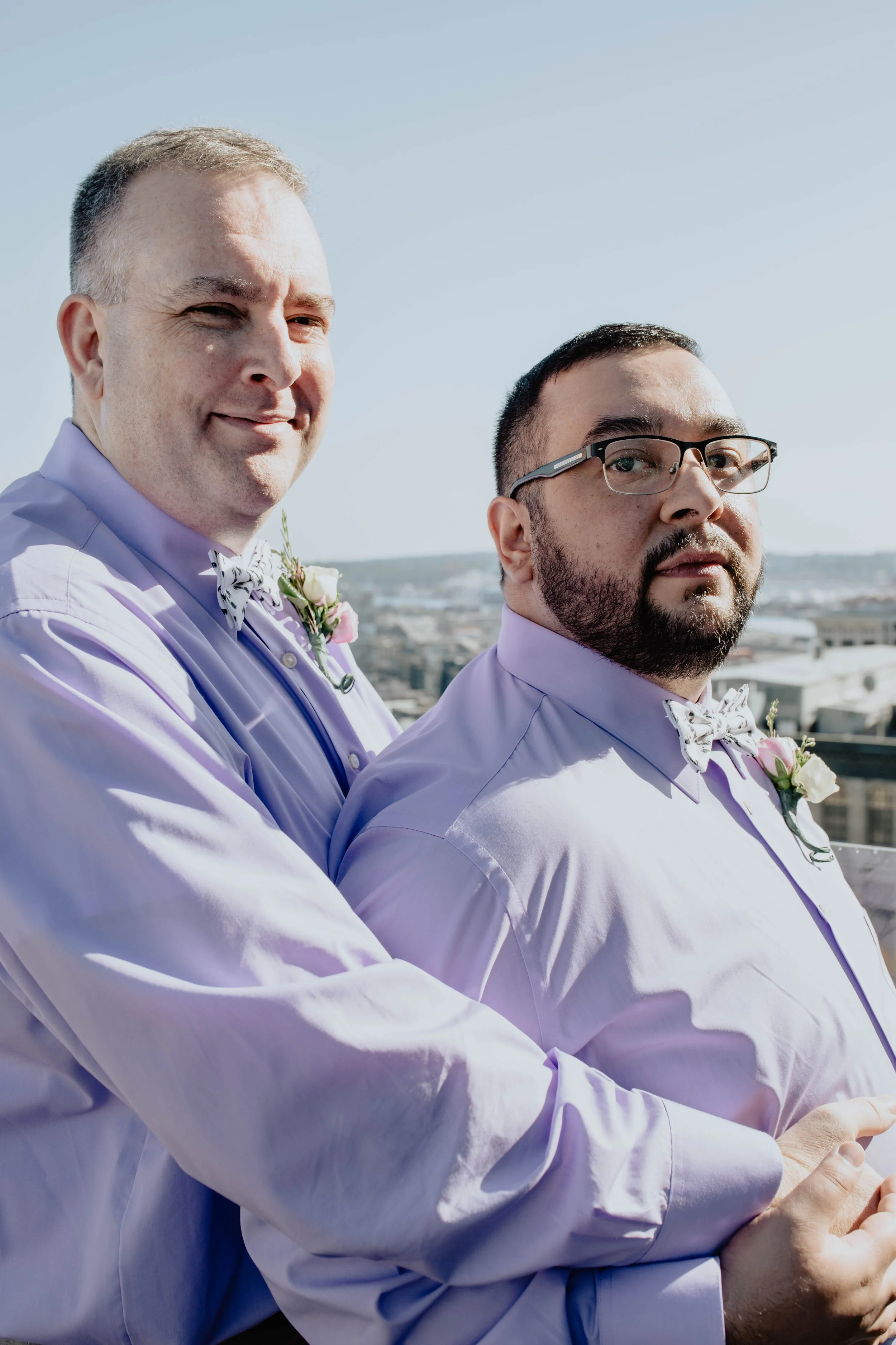 Two men wearing lavender shirts and bow ties, standing outdoors on a rooftop overlooking a cityscape, celebrating a wedding. Seattle Municipal Courthouse wedding photography.