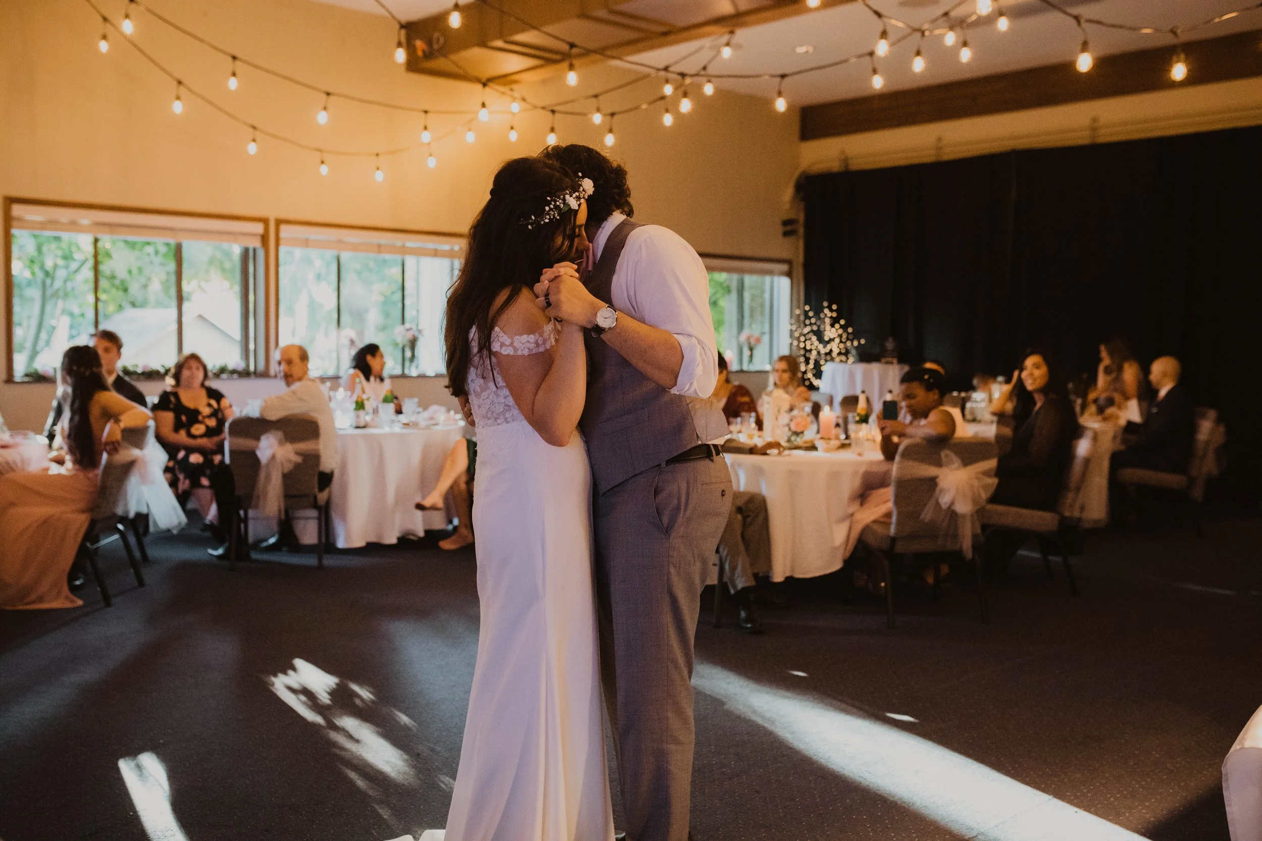 A bride and groom sharing their first dance at their wedding reception, surrounded by seated guests in a decorated banquet hall with string lights and large windows. Seattle, WA wedding photography.