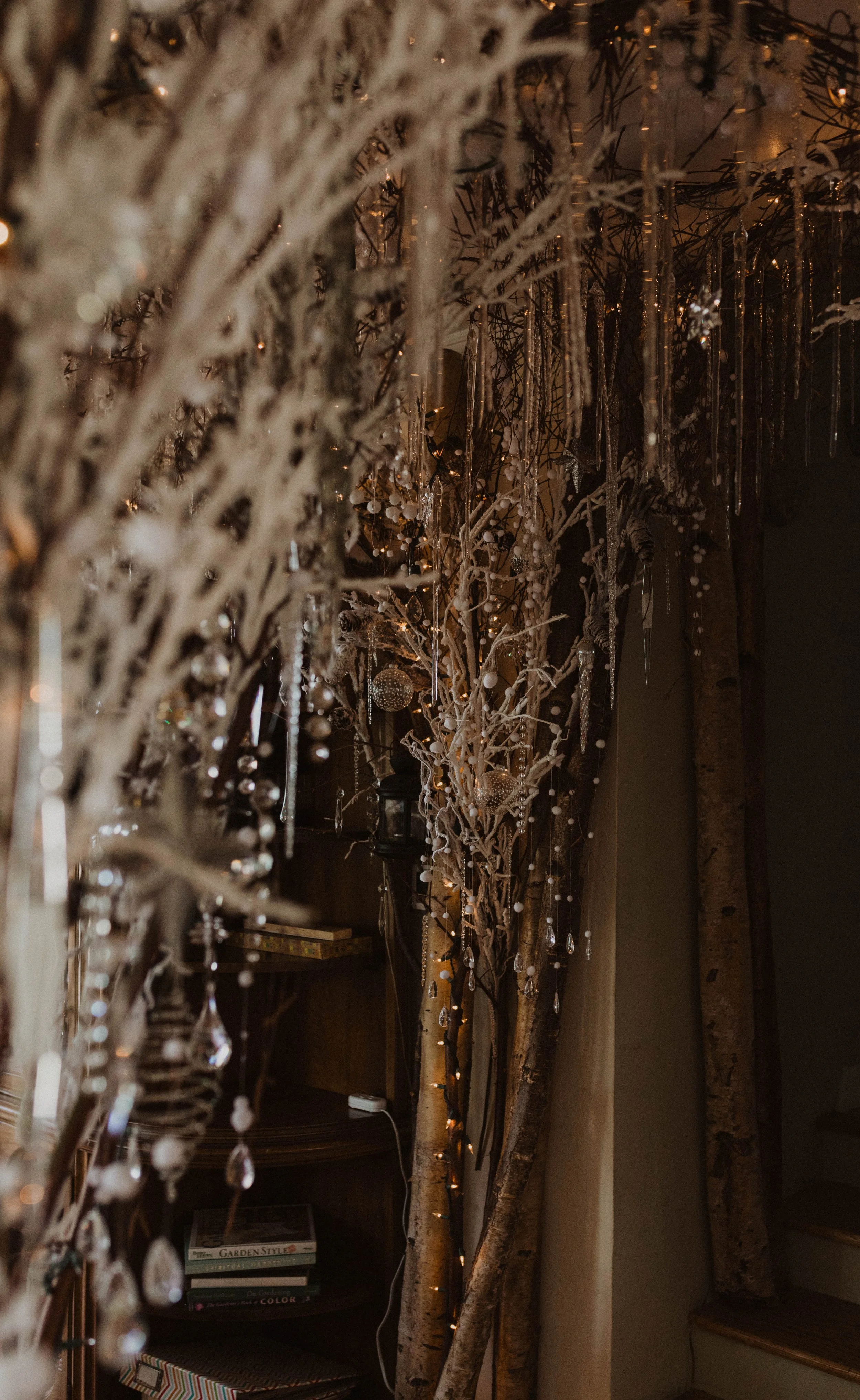 Decorated indoor space with a tall, white, leafless tree adorned with fairy lights, hanging crystal ornaments, and decorative baubles, alongside additional branches decorated in a similar style, with a dark background and a wooden staircase to the ri