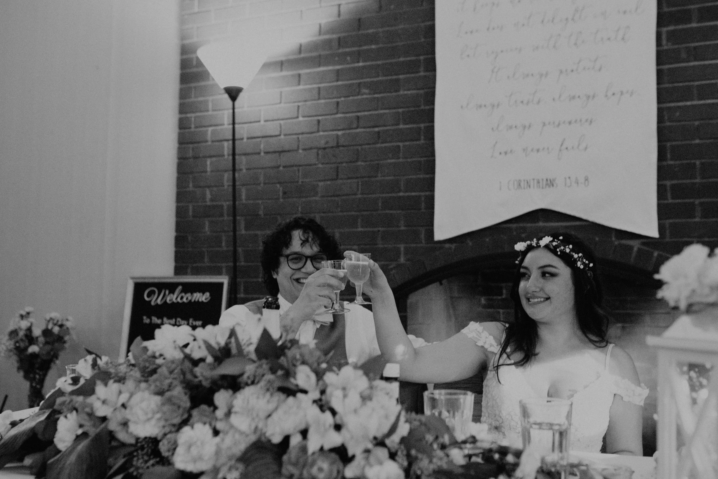A black and white photo of a wedding celebration. A man and woman, likely the bride and groom, are sitting at a table, smiling and raising glasses in a toast. The man has curly hair and glasses, and the woman is wearing a floral crown and a lace dres