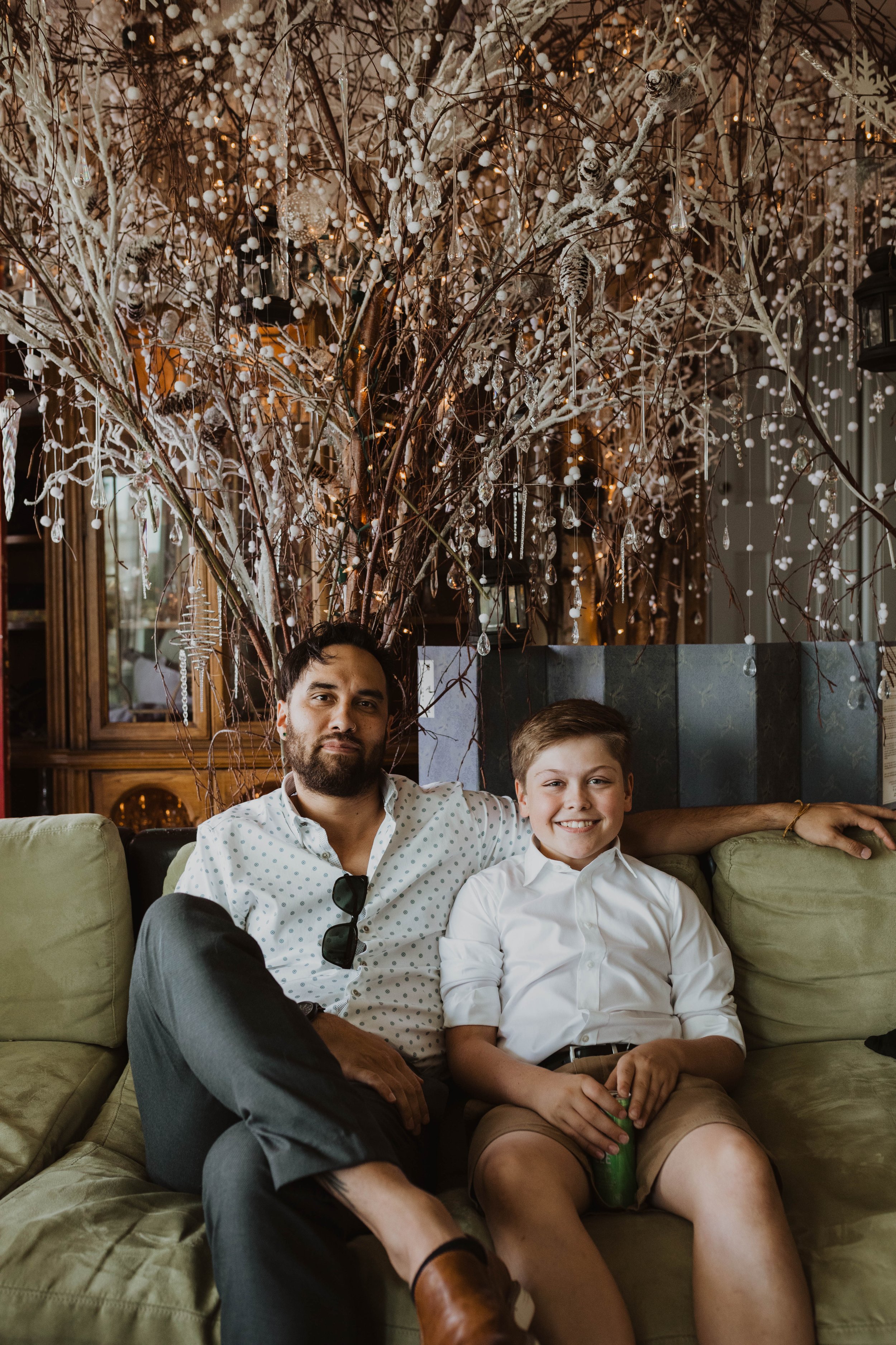 A man and a boy sitting on a couch, with a decorative tree behind them, in a warmly lit indoor setting. Seattle, WA wedding photography.