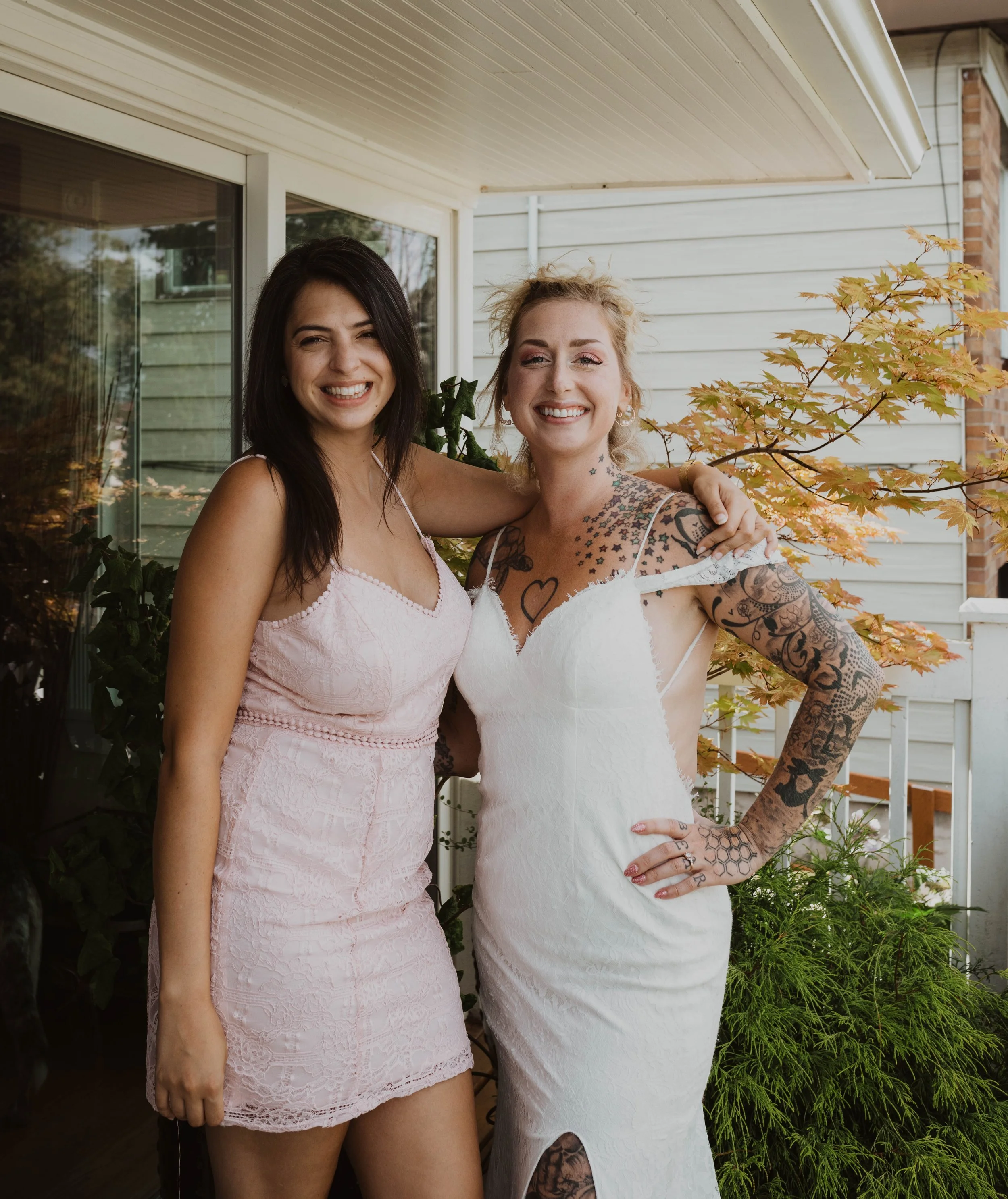 Two women smiling and posing together outside, one wearing a pink lace dress, the other in a white dress with tattoos, on a porch with plants and a tree with yellow leaves. Seattle, WA wedding photography.