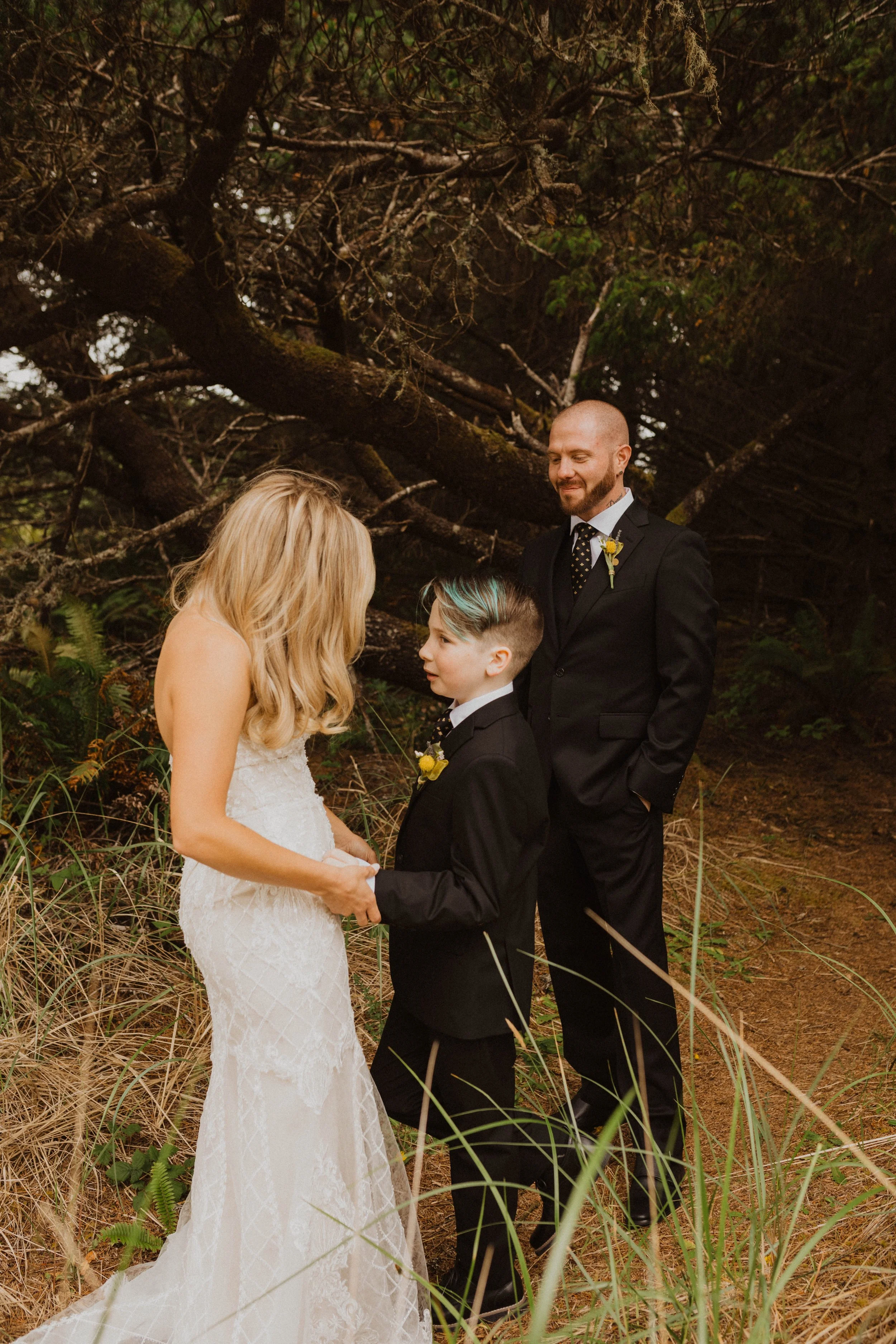 A bride and a young boy holding hands while standing in front of an officiant and a groom, outdoors in a wooded area with large trees and tall grass. Long Beach, WA wedding photography.