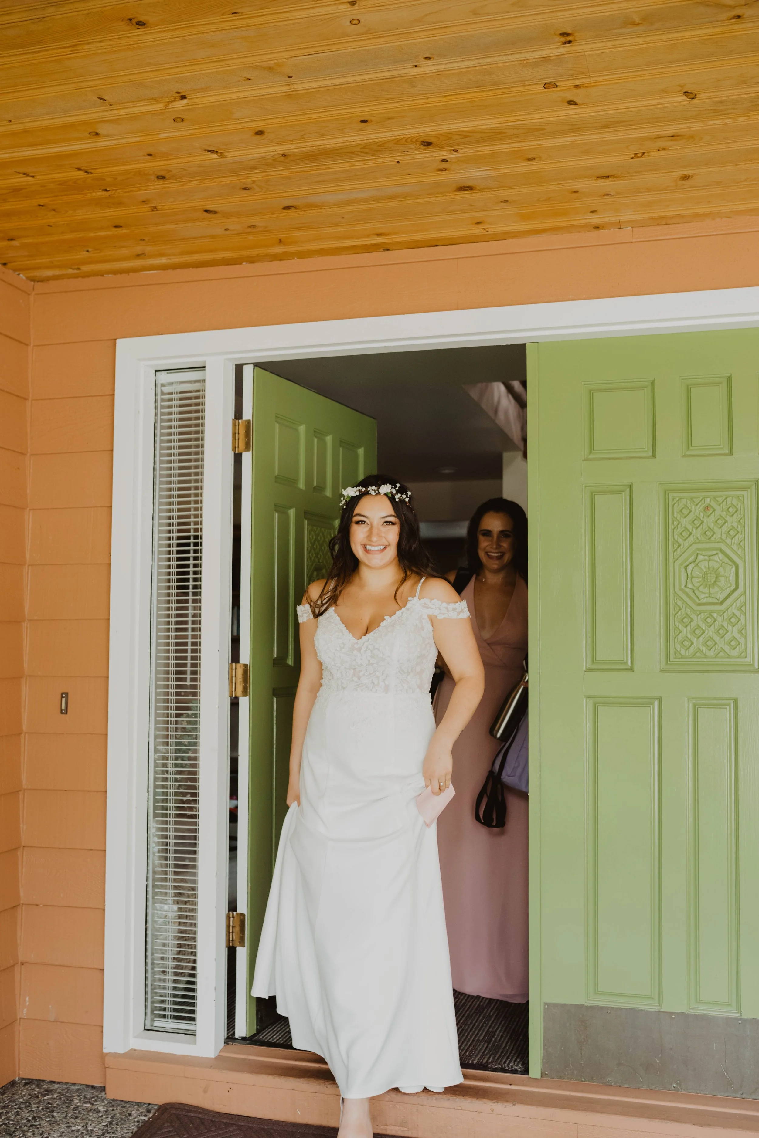 Bride in a white wedding dress smiling and walking out of a building, with a woman in a pink dress behind her, through a green door. Seattle, WA wedding photography.
