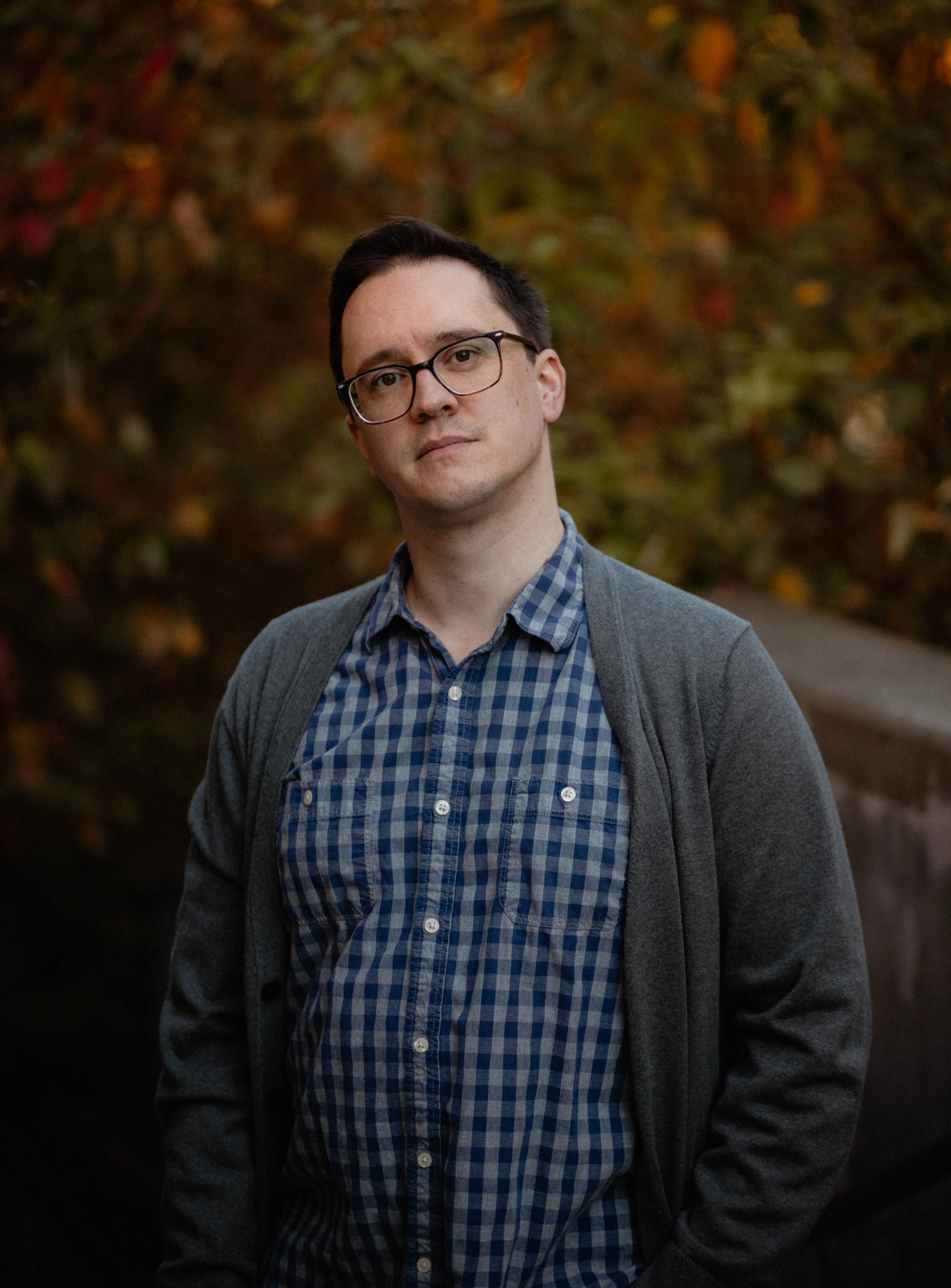 A young man with glasses, wearing a blue checkered shirt and gray cardigan, standing outdoors in front of fall foliage. Seattle professional head shot photography