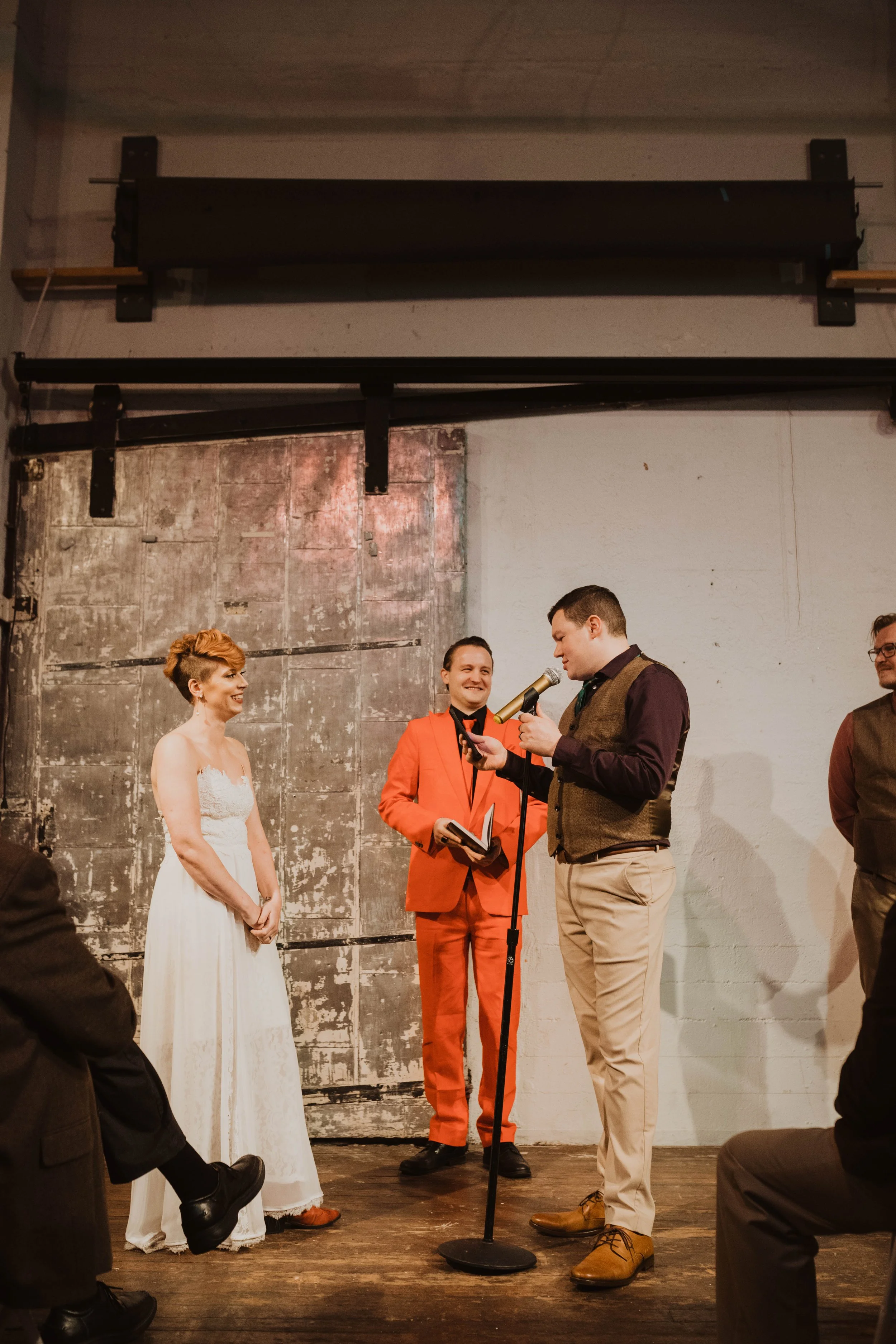 A wedding ceremony taking place indoors with four people, including a bride in a white dress, a groom in a brown vest and light pants, a person in an orange suit, and another person in a brown vest; the groom is speaking into a microphone while holdi