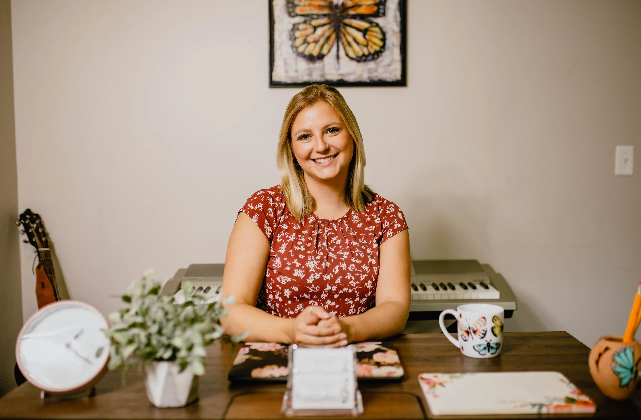 A woman with blonde hair wearing a floral red shirt sits at a wooden desk with a smile. On the desk are a potted plant, a decorative clock, a colorful butterfly mug, and a notebook or planner. In the background, there is a digital keyboard and a fram