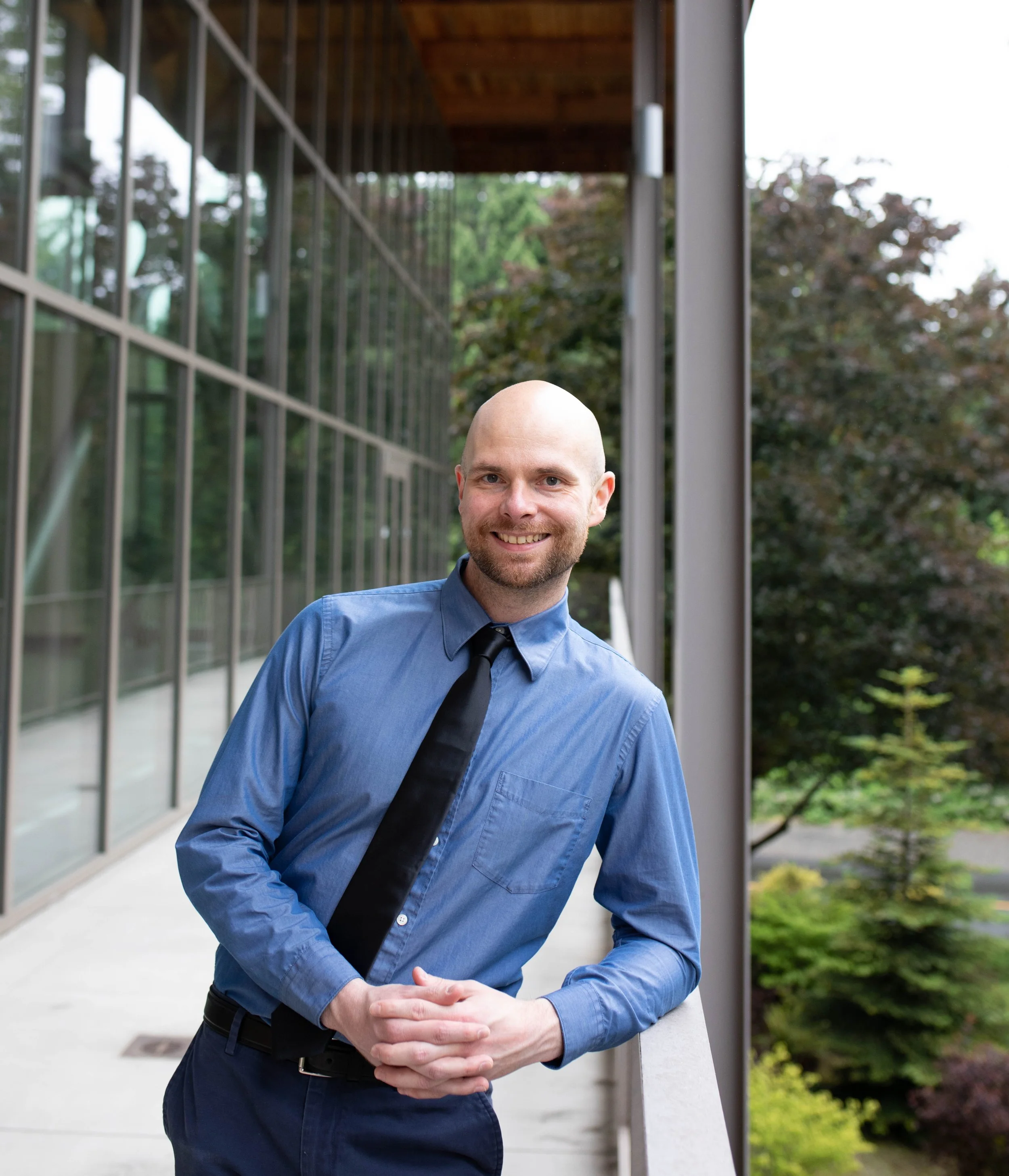 A smiling man with a shaved head and beard, dressed in a blue shirt and black tie, leaning on a balcony railing outdoors with modern glass building and trees in the background. Seattle professional head shot photography