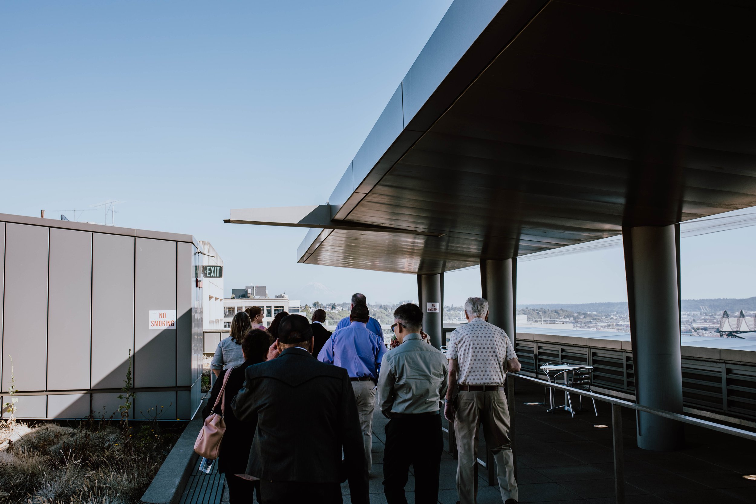 A group of people standing and walking on a rooftop or balcony area with modern architecture, under a large overhang, with a city skyline in the background. Seattle Municipal Courthouse wedding photography.