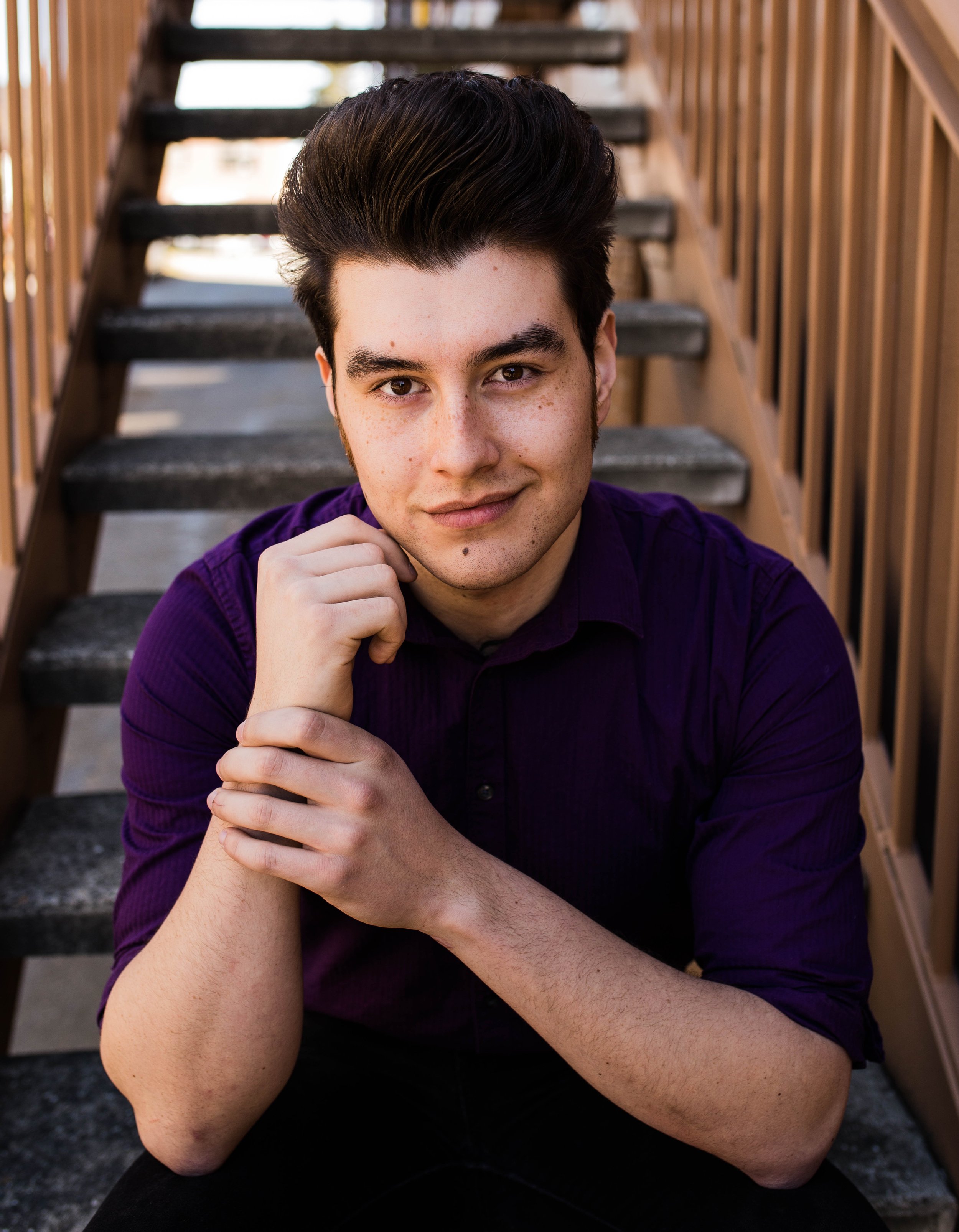 A young man with dark hair and a slight smile, wearing a purple shirt, sitting on outdoor stairs with wooden railings, resting his chin on his hand. Seattle professional head shot photography