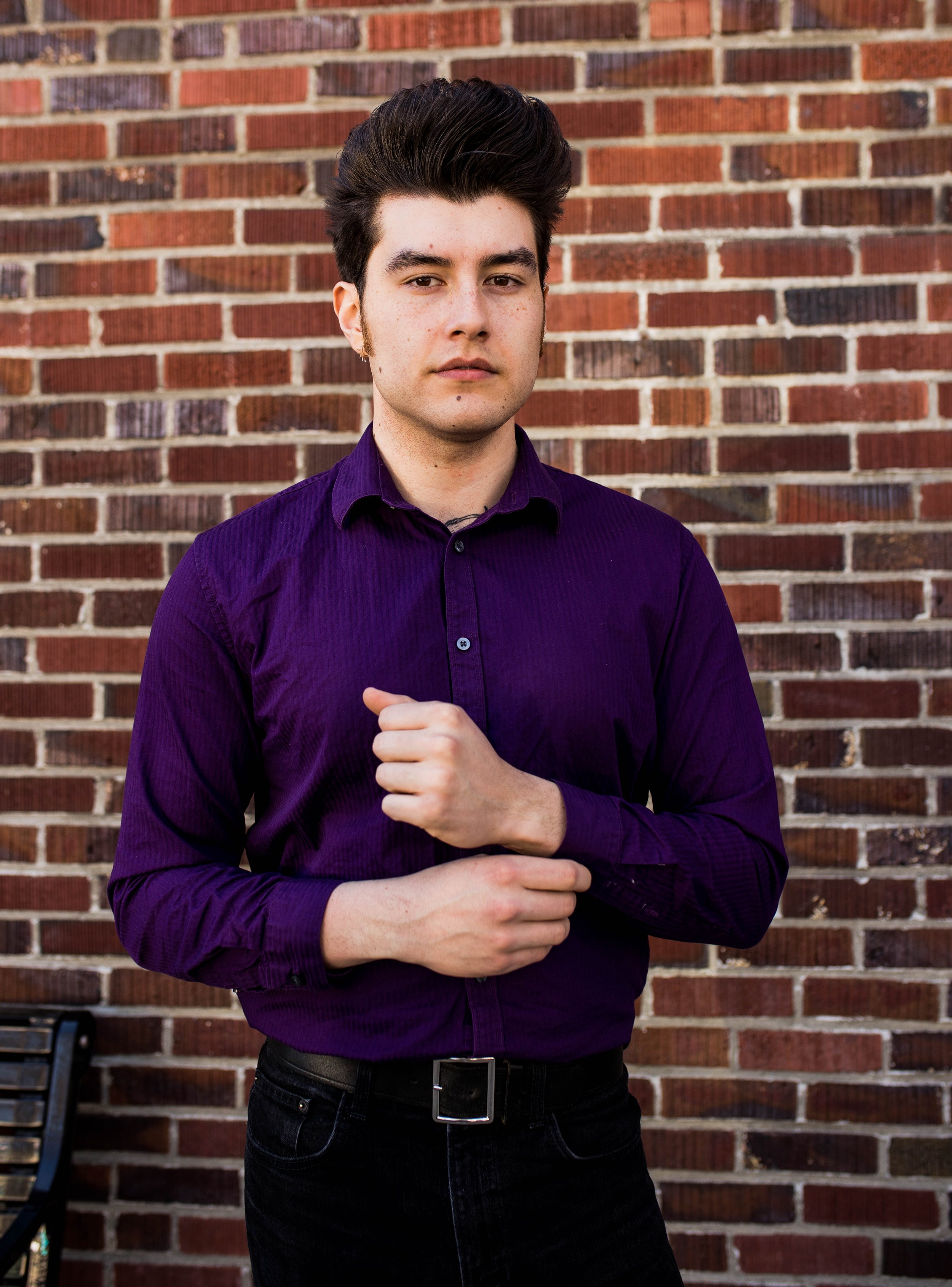 A young man with dark hair is standing in front of a brick wall, wearing a purple shirt and black pants, looking at the camera with a serious expression. Seattle professional head shot photography