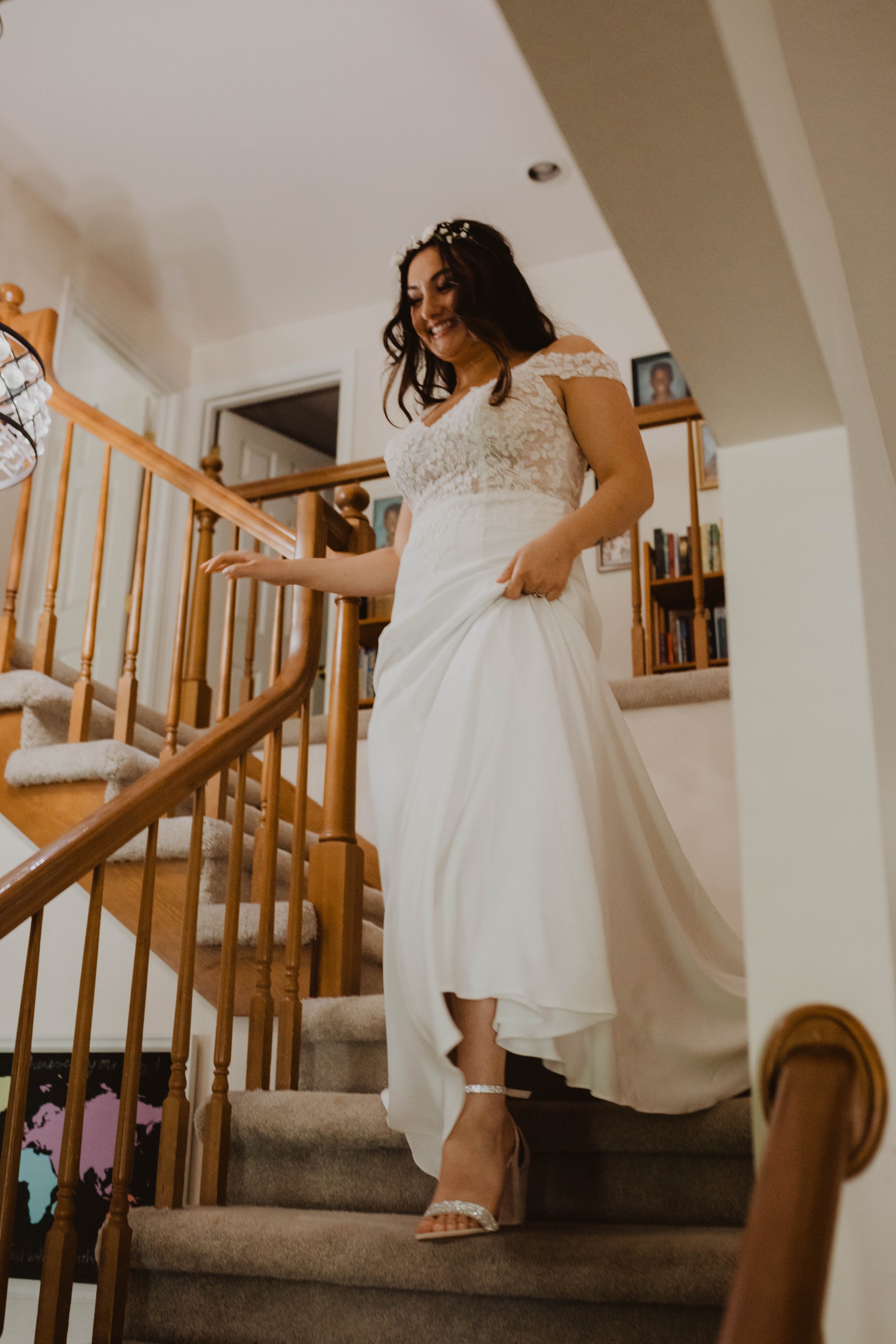 A bride in a white lace wedding dress and high heels descending a carpeted staircase inside a house, smiling. Seattle, WA wedding photography.