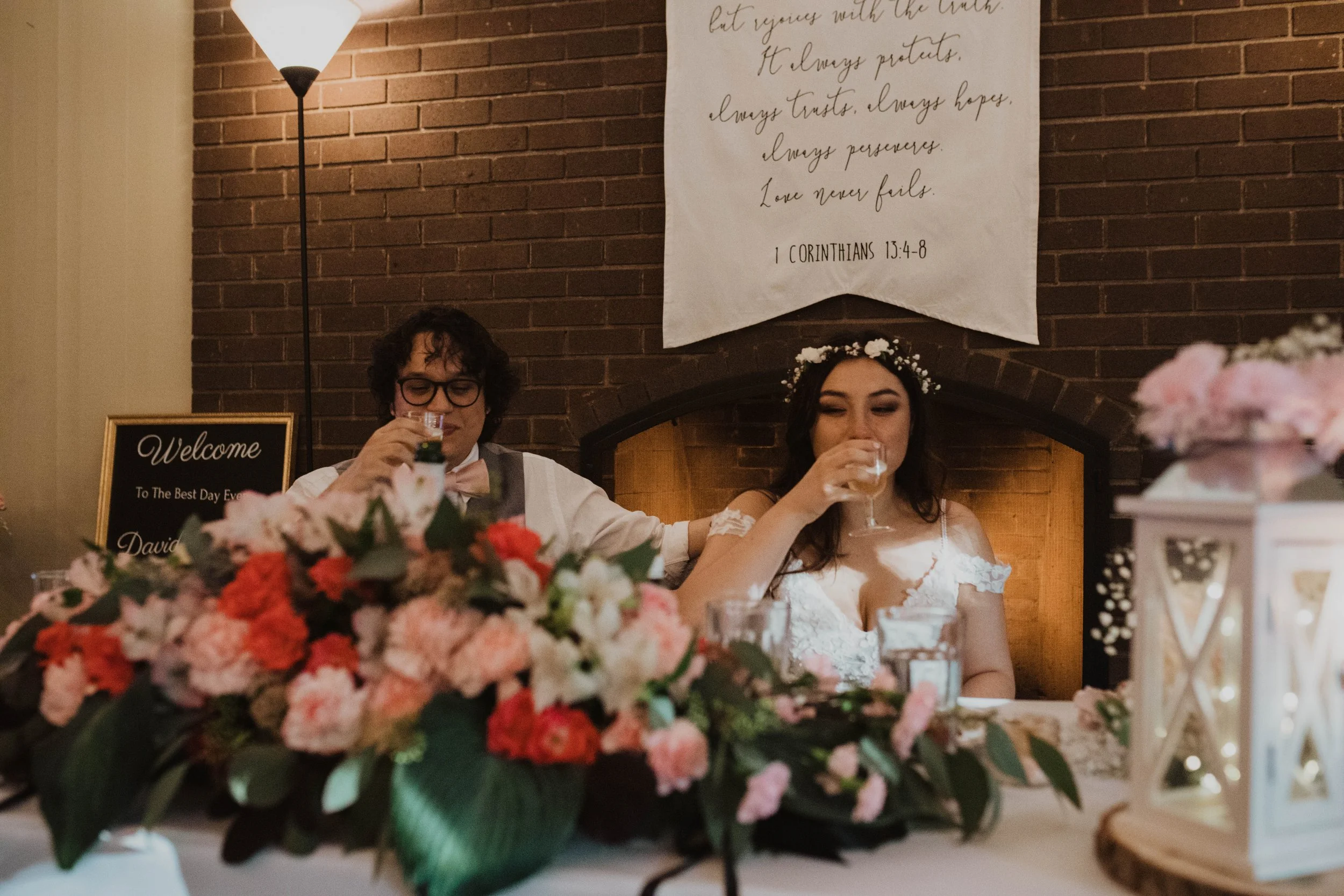 Bride and groom sitting at a wedding reception table, drinking from glasses, with a floral centerpiece and lanterns, brick wall with a wedding quote banner in the background. Seattle, WA wedding photography.