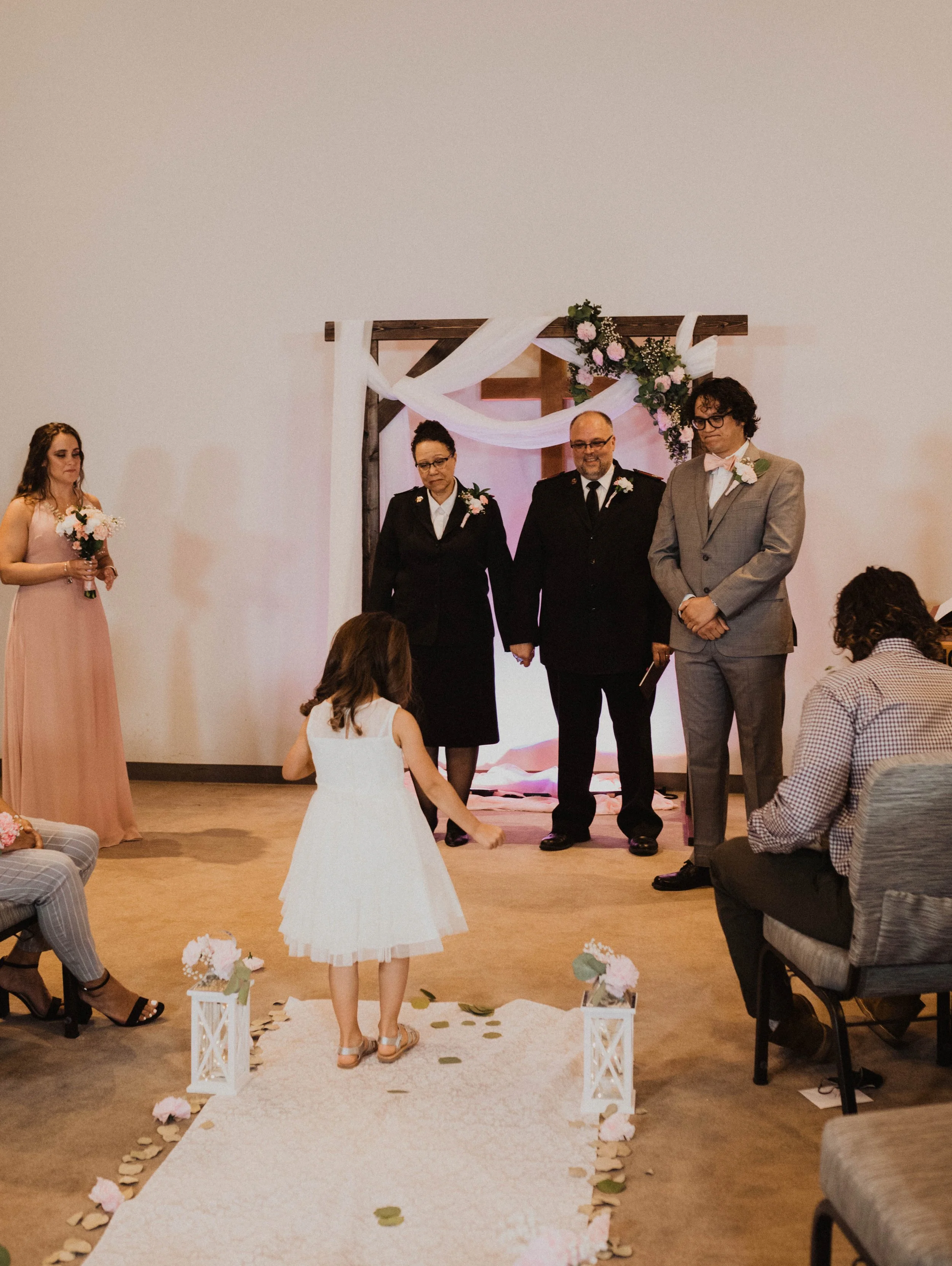 A young girl in a white dress throws flower petals during a wedding ceremony in front of the officiants and the wedding party, with floral decorations and a draped fabric backdrop. Seattle, WA wedding photography.