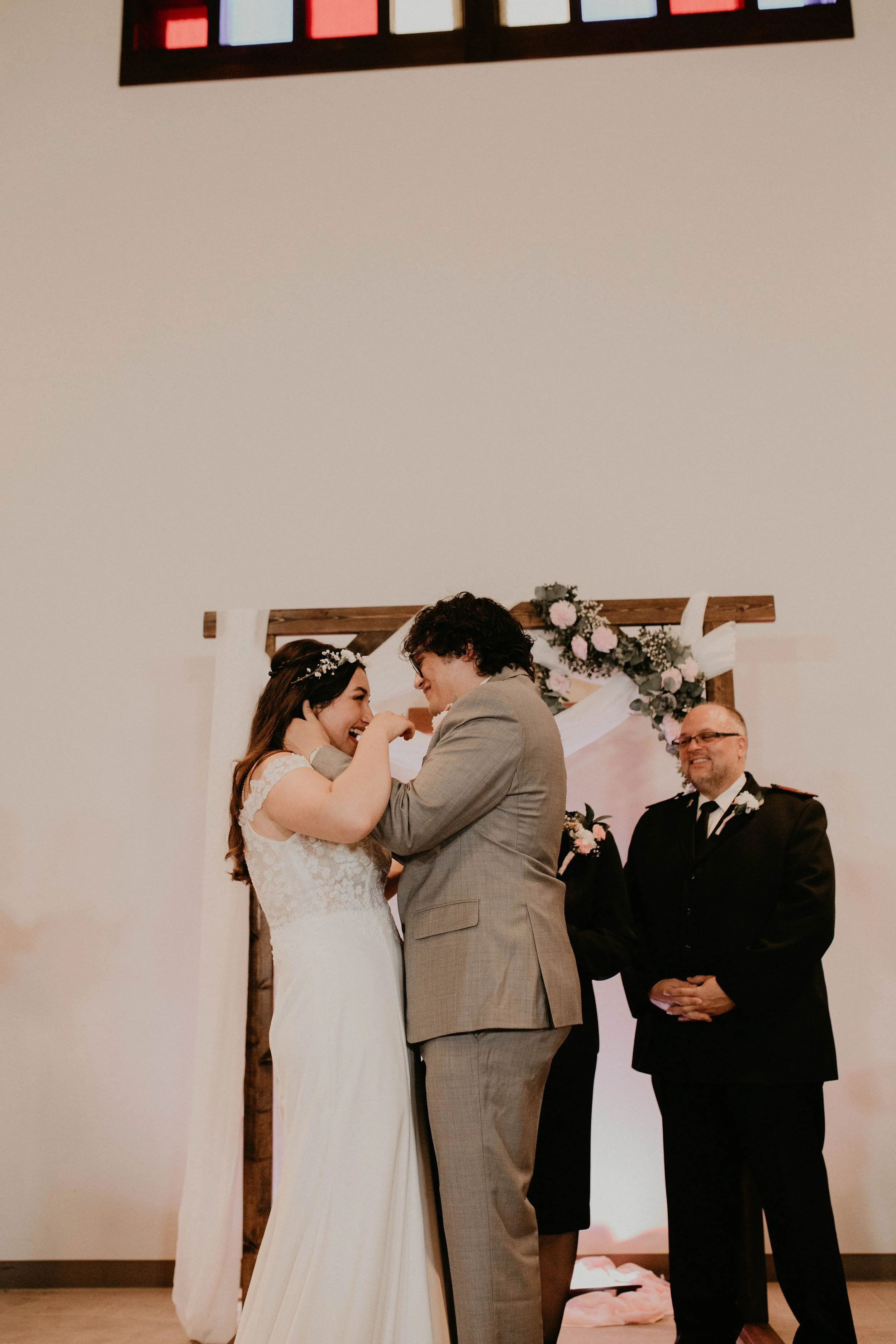 A bride and groom stand close together during their wedding ceremony, smiling and touching foreheads, with an officiant and a man in uniform nearby, under a decorated floral arch Seattle, WA wedding photography.