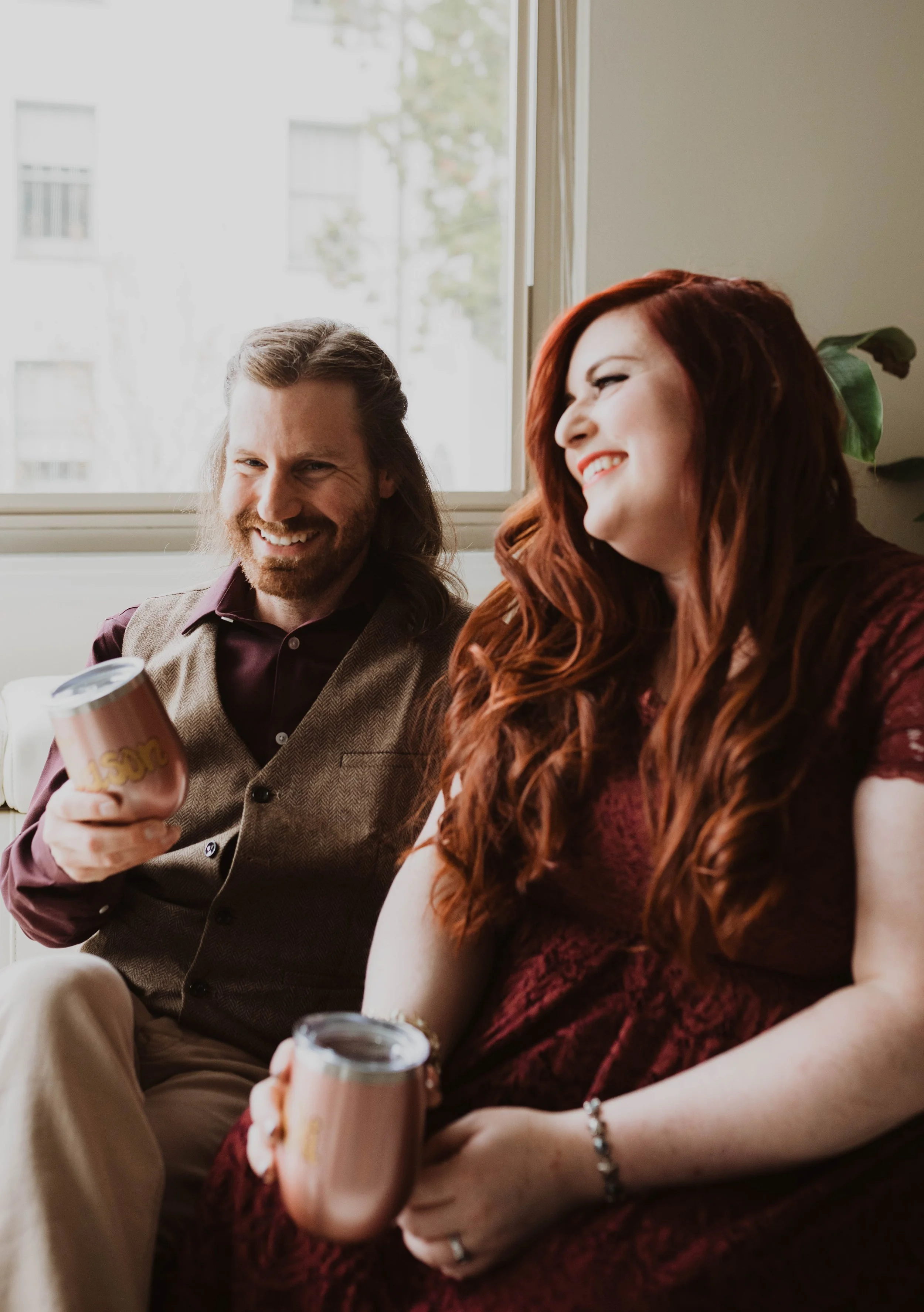 Two smiling people, a man with long hair and beard in a vest, and a woman with long red hair, sitting on a couch by a window, holding mugs and enjoying conversation. Pioneer Square, Seattle, WA wedding photography.