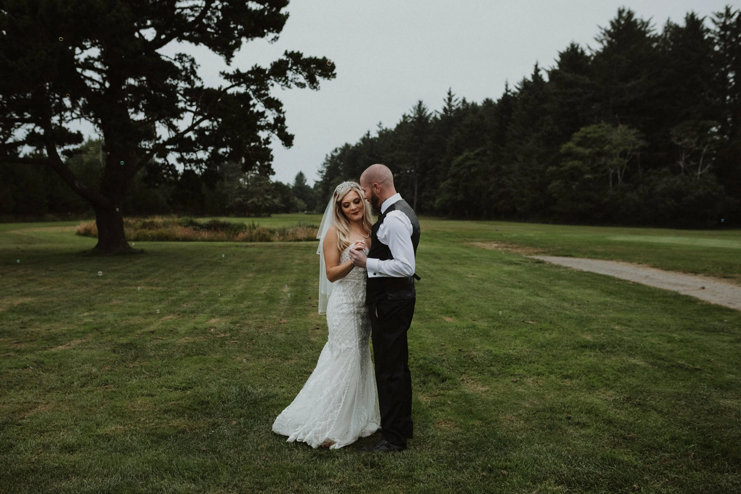 A bride and groom dance together on a grassy field during their outdoor wedding, surrounded by trees. Long Beach, WA wedding photography.