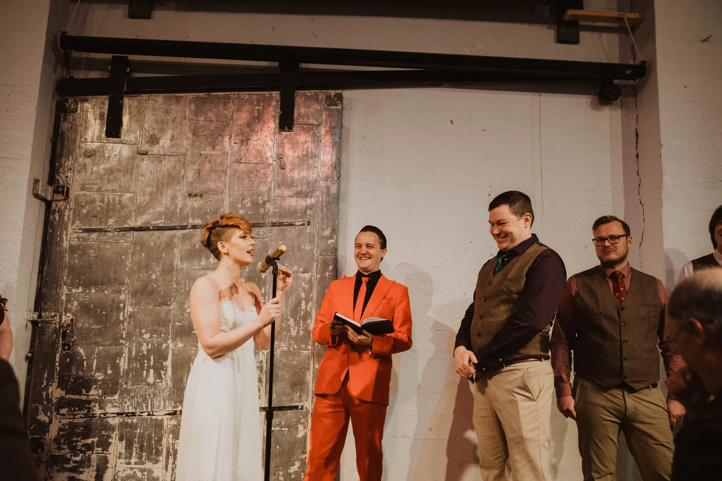 A woman in a white dress speaking into a microphone during a wedding ceremony, with three men in formal attire standing nearby, smiling and listening, in front of an industrial-style backdrop. Pioneer Square, Seattle, WA wedding photography.