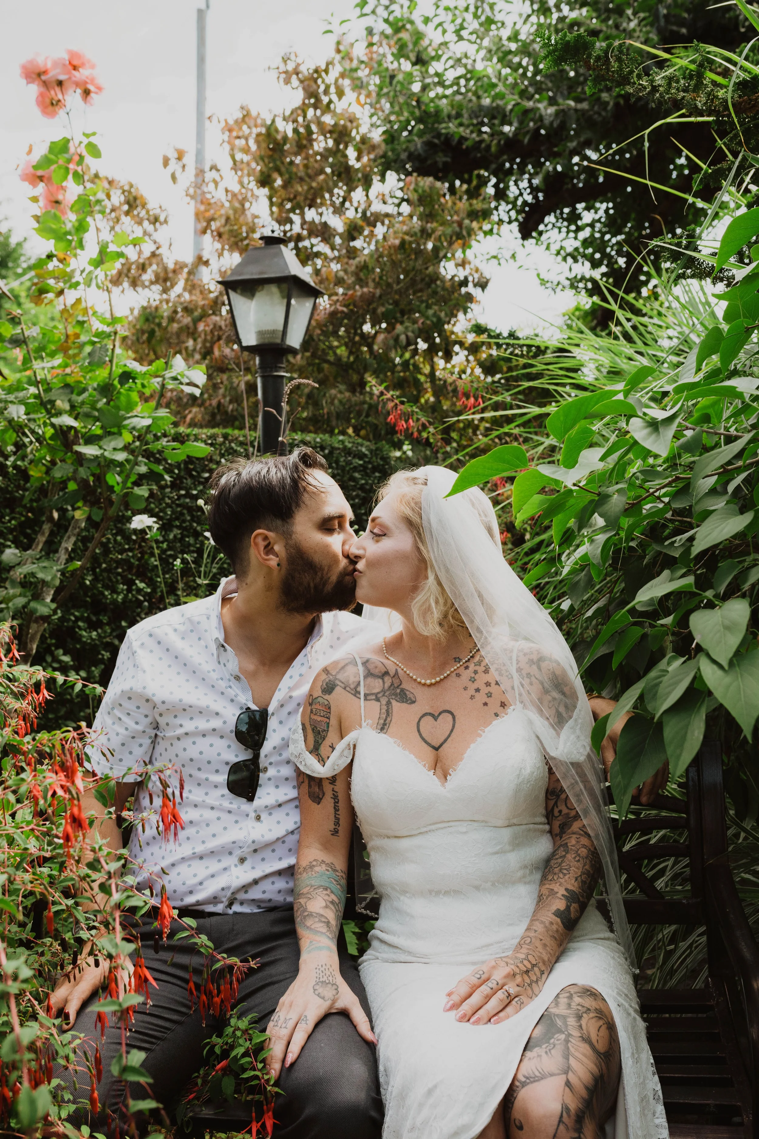 A couple sharing a kiss outdoors, surrounded by green plants and bushes, with a vintage street lamp in the background. Seattle, WA wedding photography.