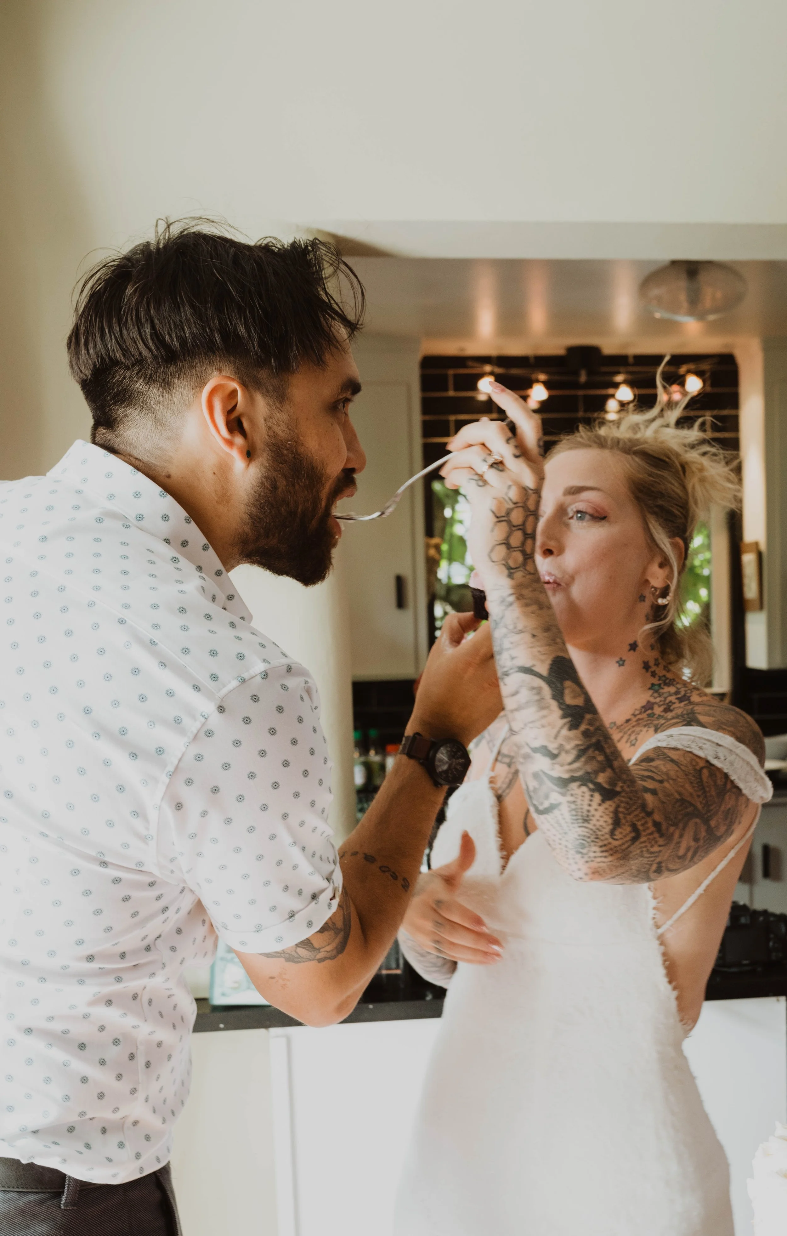 A man and a woman with tattoos share a playful moment indoors, with the man holding a spoon near the woman's mouth. Seattle, WA wedding photography.
